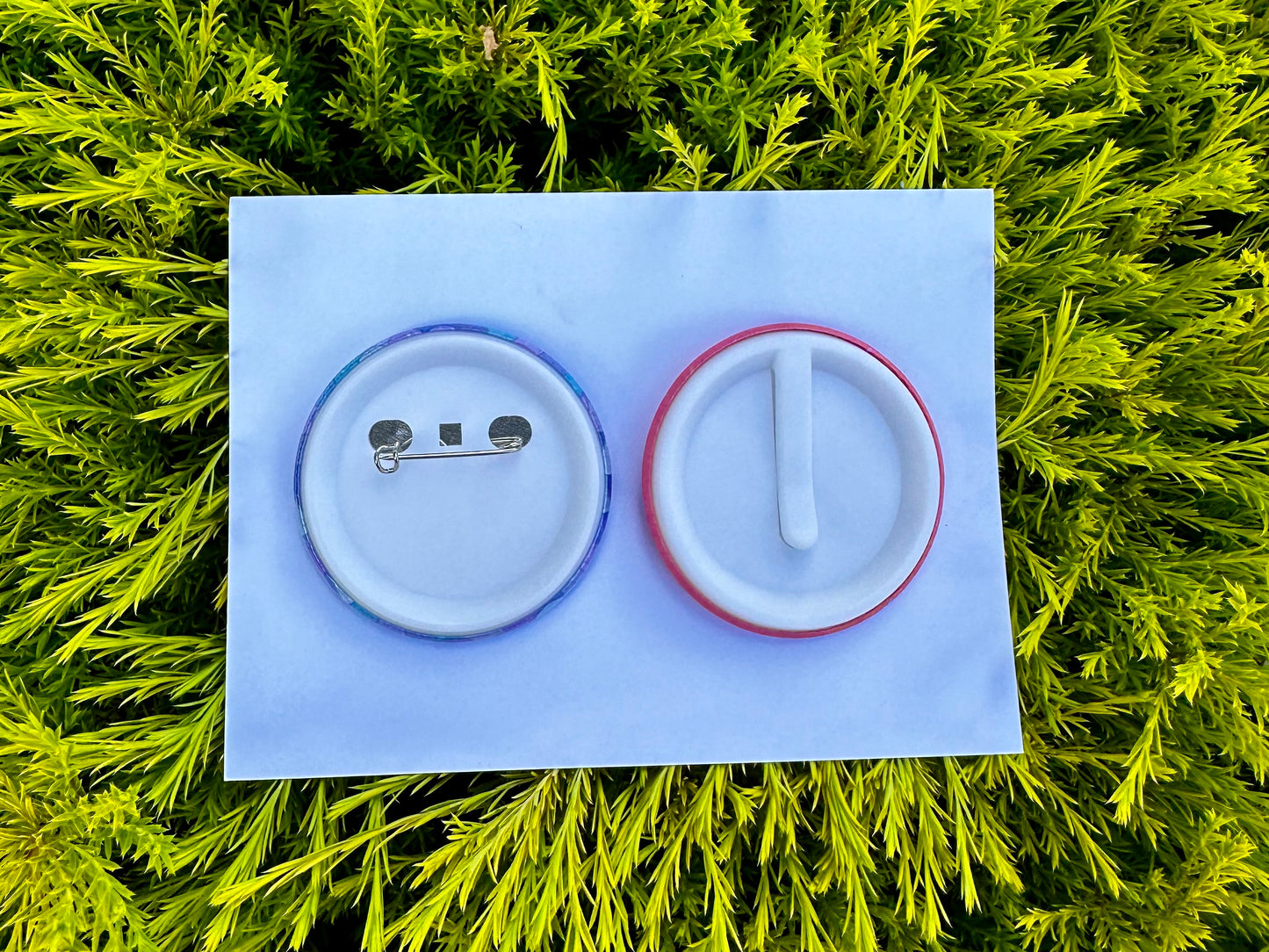 Two backs of a birthday badge on a white card, laying on green leaves. The first backing option is a standard pin backing using a safety pin. The second option is a safety slider which clips and slides over documents, clothing and material.