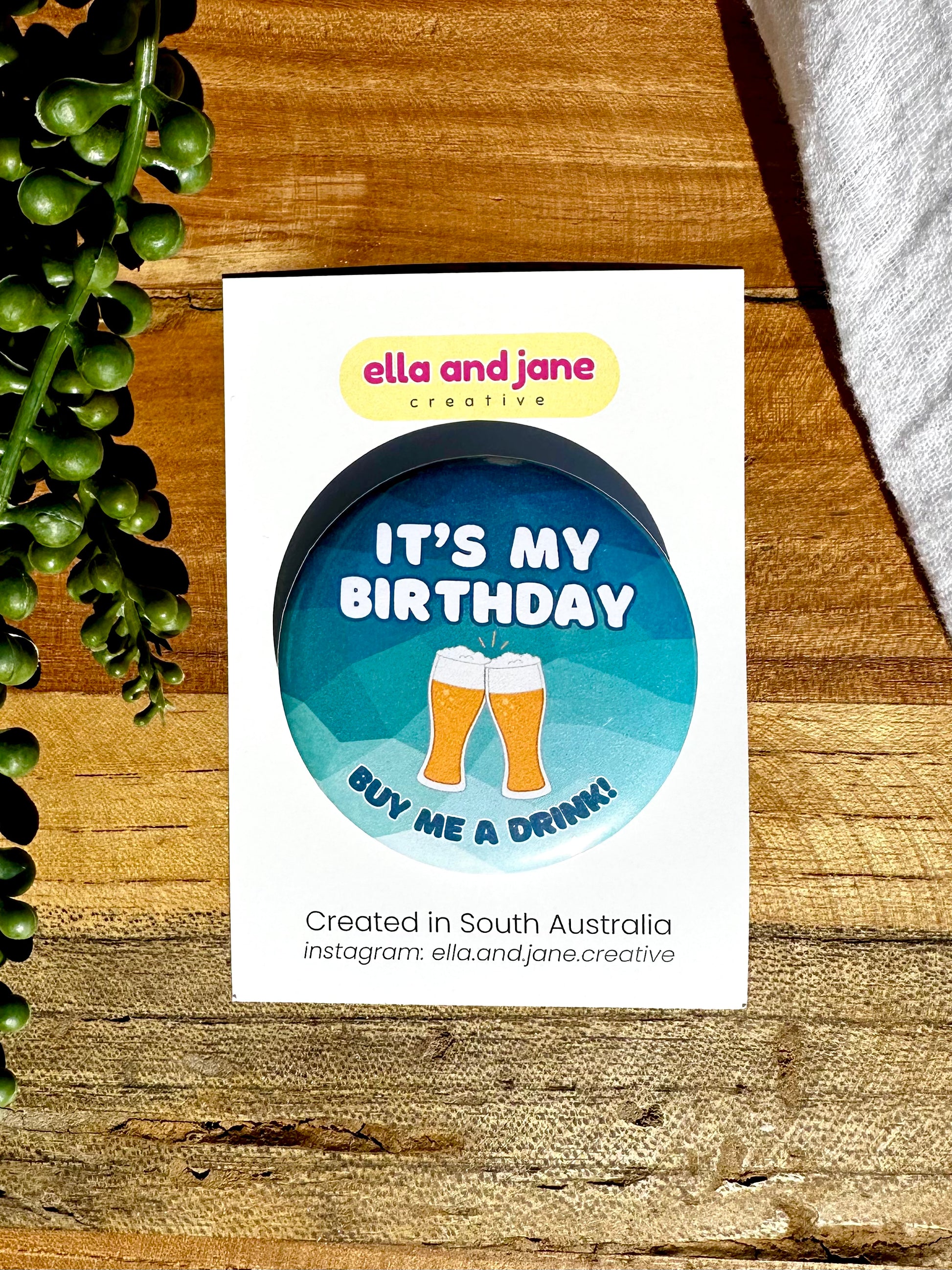 A blue and green, round birthday badge with It's My Birthday Buy Me A Drink written as text and with a picture of two beer glasses filled with beer touching each other to give a cheers. The party badge is sitting on a product card and laying on a piece of wood with decorations.