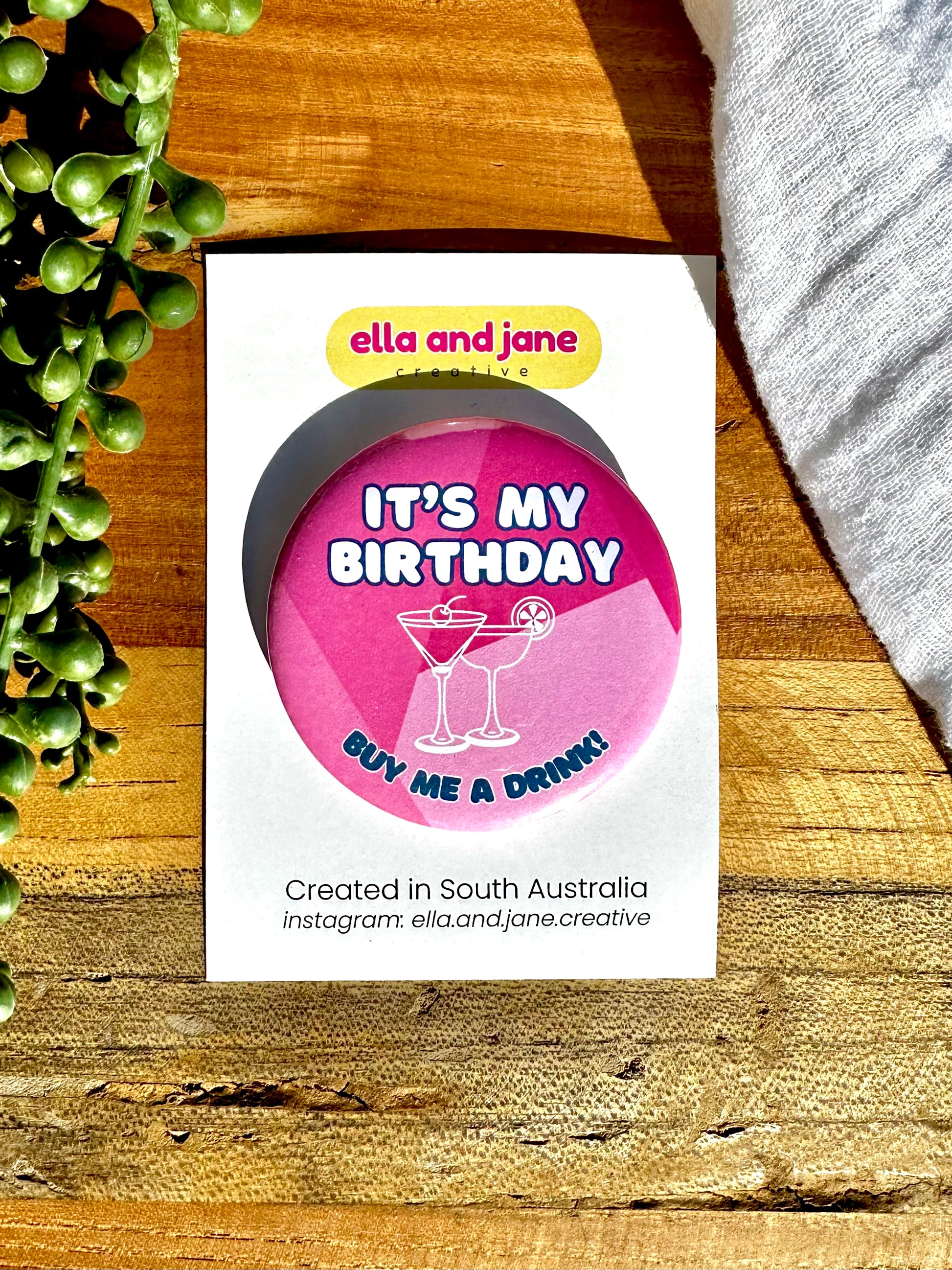 A pink, round birthday badge with It's My Birthday Buy Me A Drink written as text and with a picture of two cocktail glasses a martini and margarita. The party badge is sitting on a product card and laying on a piece of wood with decorations.