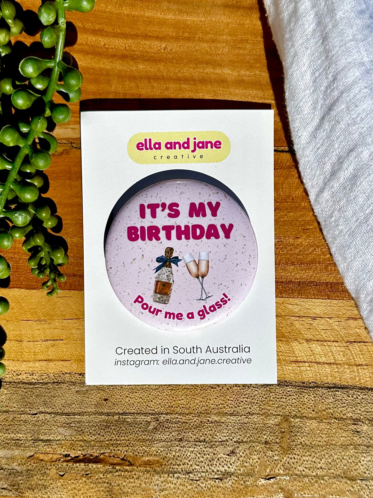 A pink, round birthday badge with It's My Birthday Pour Me A Glass written as text and with a picture of champagne glasses and sparkling wine bottle with a blue bow attached. The badge is sitting on a product card and laying on a piece of wood with decorations.