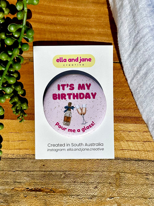 A pink, round birthday badge with It's My Birthday Pour Me A Glass written as text and with a picture of champagne glasses and sparkling wine bottle with a blue bow attached. The badge is sitting on a product card and laying on a piece of wood with decorations.