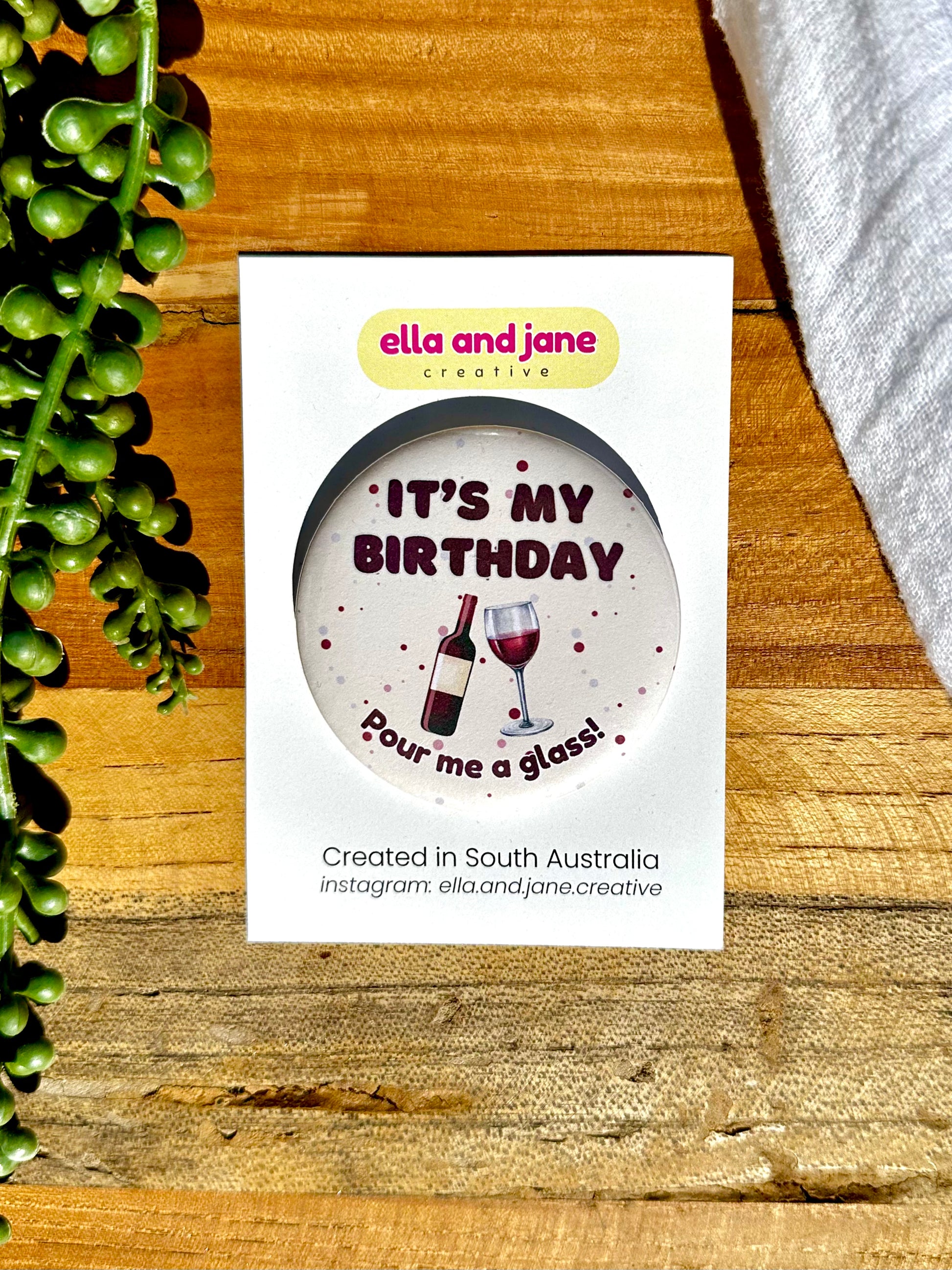 A cream, round birthday badge with It's My Birthday Pour Me A Glass written as text and with a picture of a red wine glass and a red wine bottle. The badge is sitting on a product card and laying on a piece of wood with decorations.
