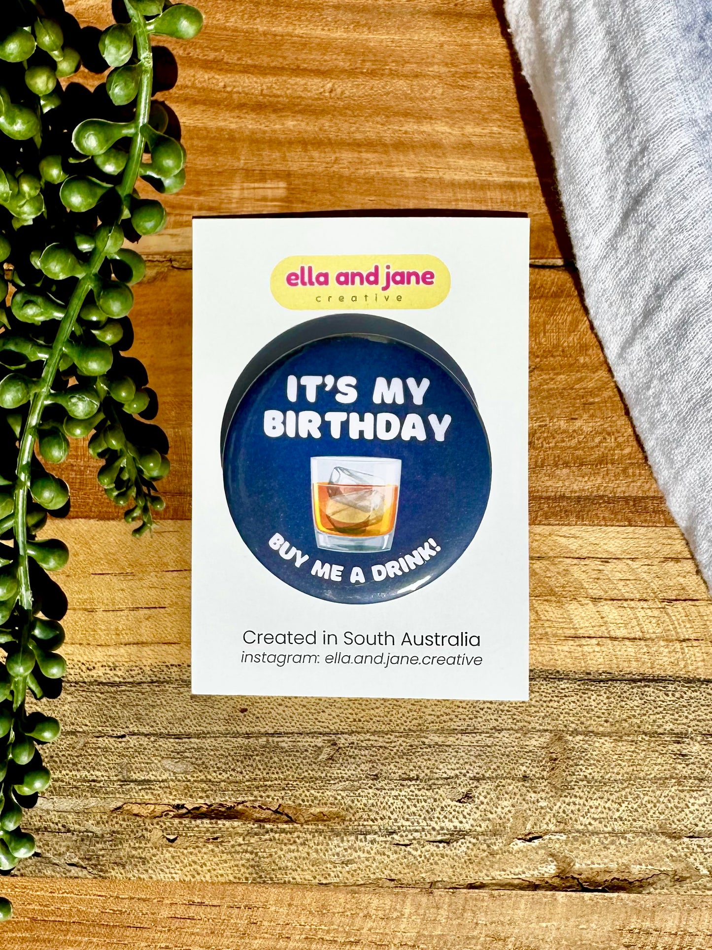 A navy blue, round birthday badge with It's My Birthday Buy Me A Drink written as text and with a picture of spirits glass filled with alcohol and ice. The badge is sitting on a product card and laying on a piece of wood with decorations.