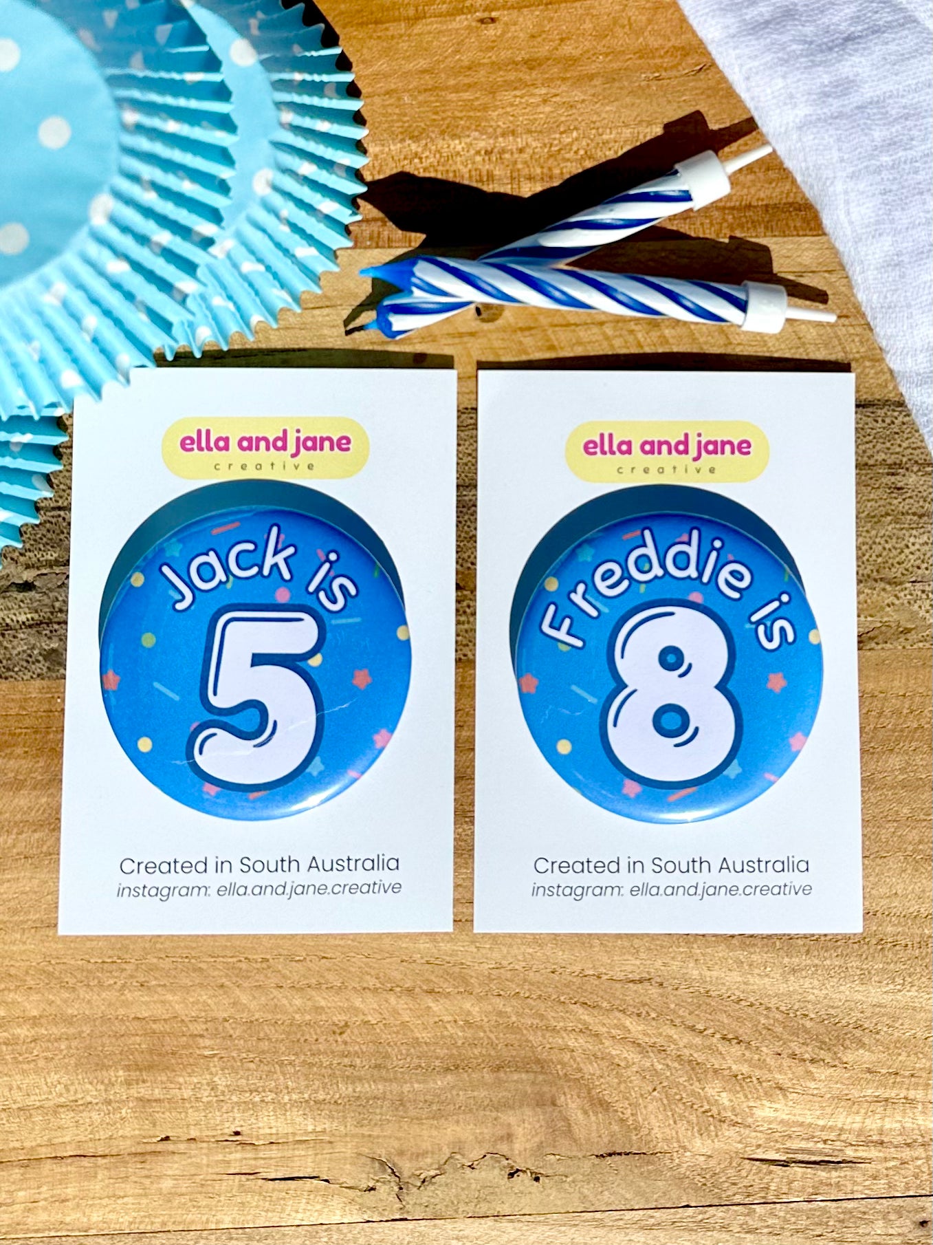 Blue birthday badges with sprinkles as the background. They are medium blue with white and blue text. One reading 'Jack is 5' and the other 'Freddie is 8'. The badges are round and are sitting on a white product card. There are birthday party supplies as decorations, including cupcake cases and birthday candles.