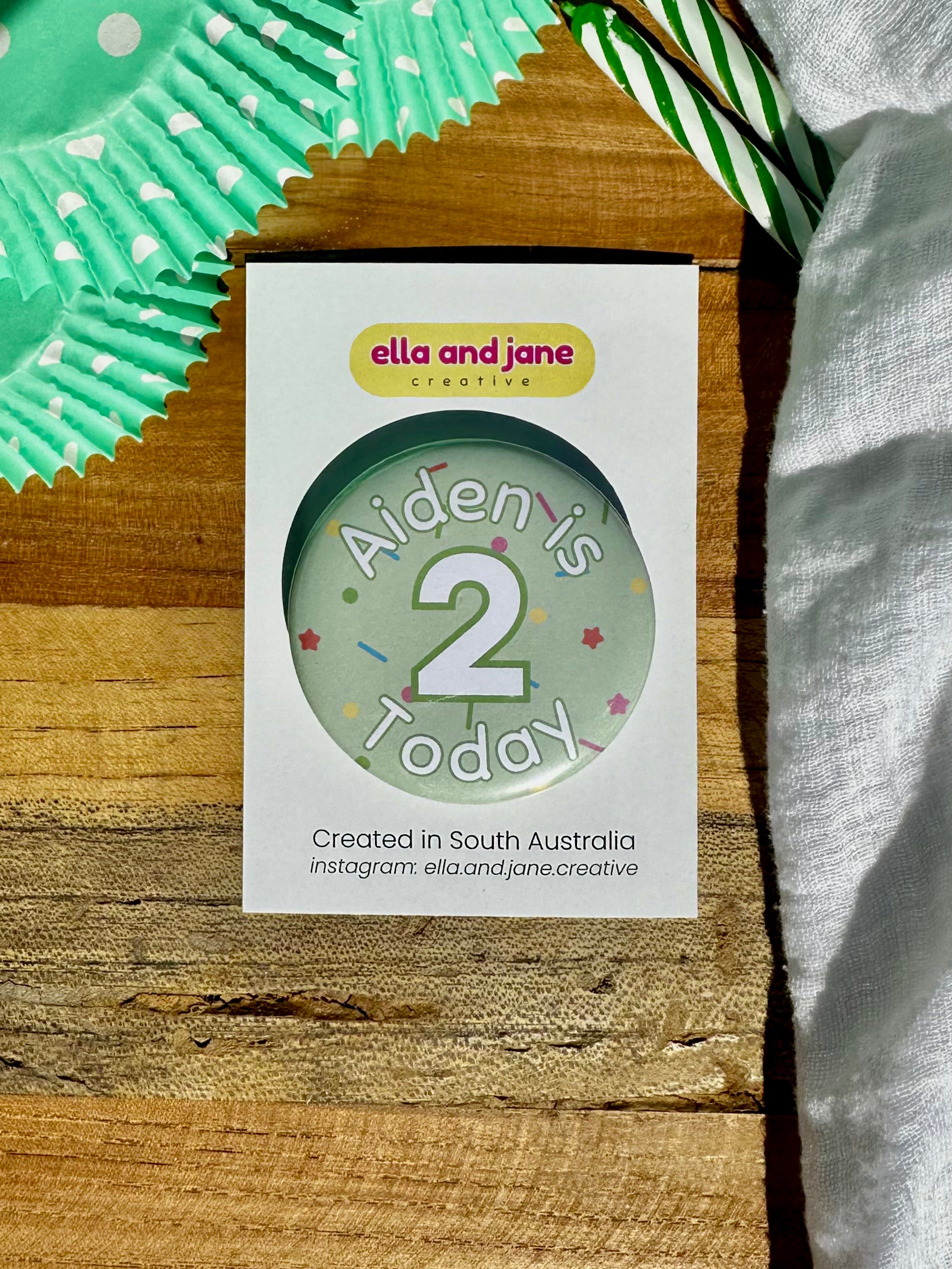 A light green birthday badge with sprinkles as the background. The text says 'Aiden is 2 Today' in white and green. The badge is round and is sitting on a white product card. There are birthday party supplies as decorations, including cupcake cases and birthday candles.
