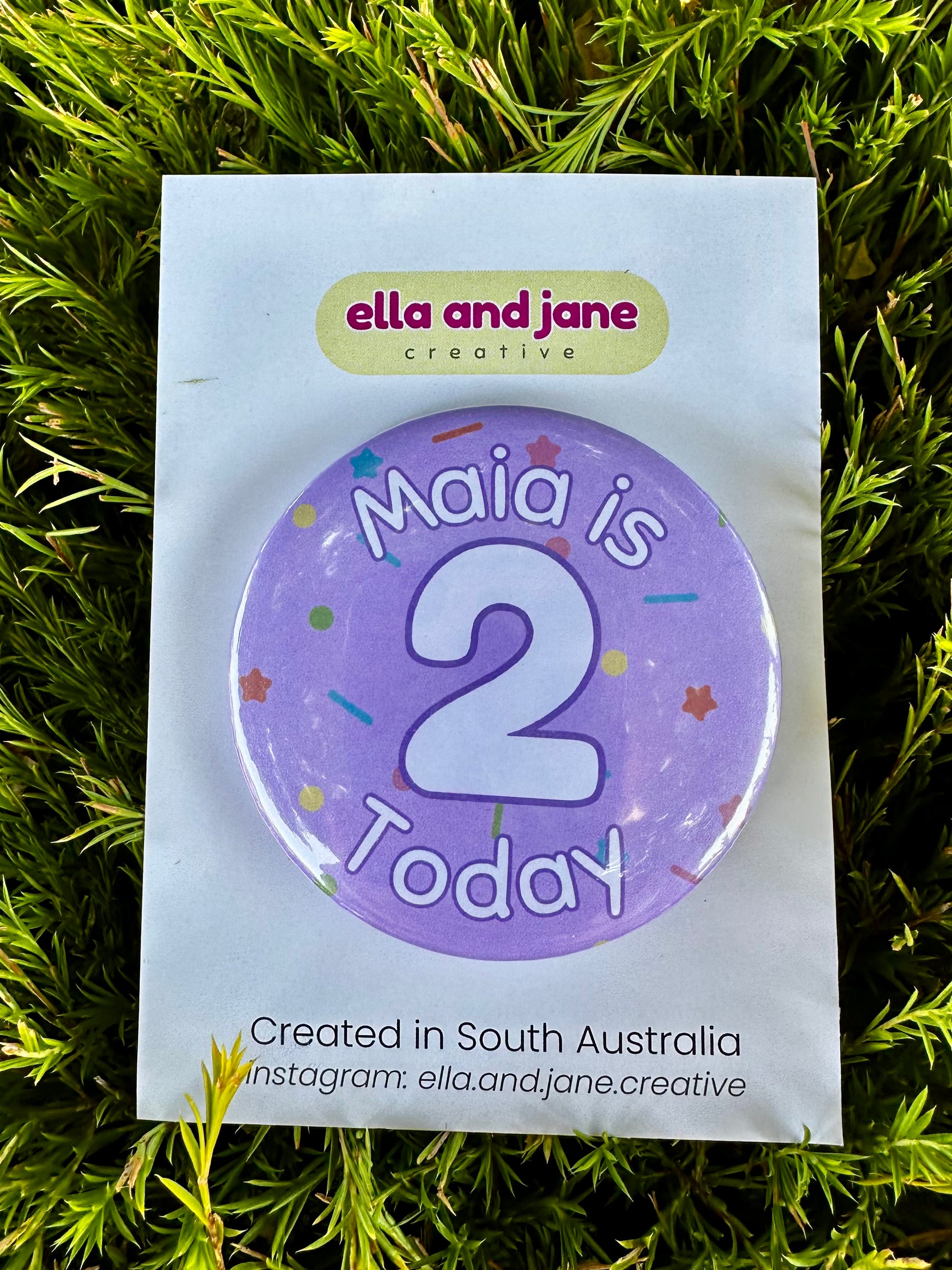 A lilac purple birthday badge with sprinkles as the background. The text says 'Maia is 2 Today' in white and purple. The badge is round and is sitting on a white product card.