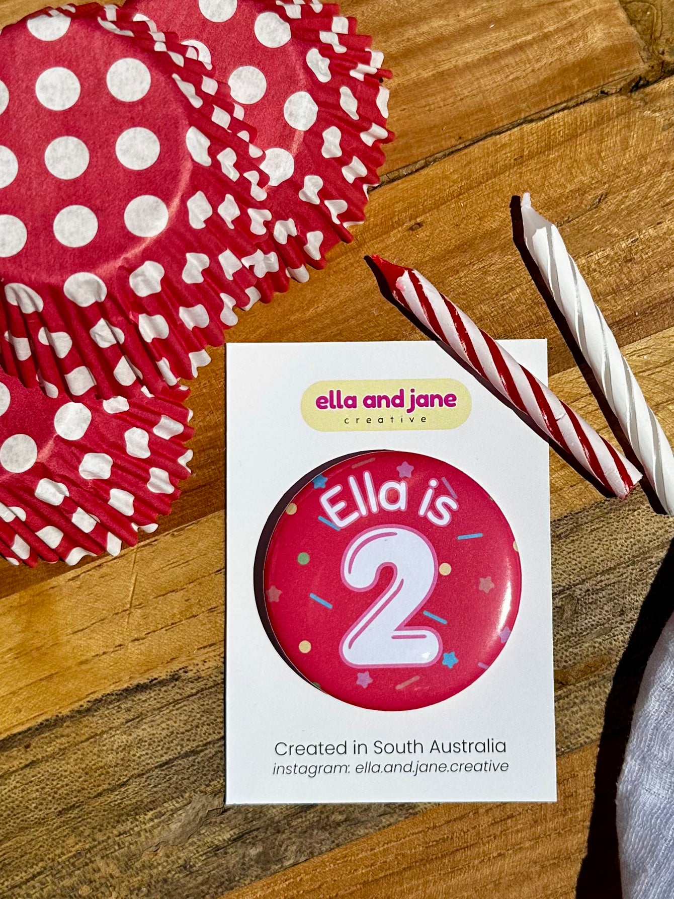 A red birthday badge with sprinkles as the background. The text says 'Ella is 2' in white and red. The badge is round and is sitting on a white product card. There are birthday party supplies as decorations, including cupcake cases and birthday candles.