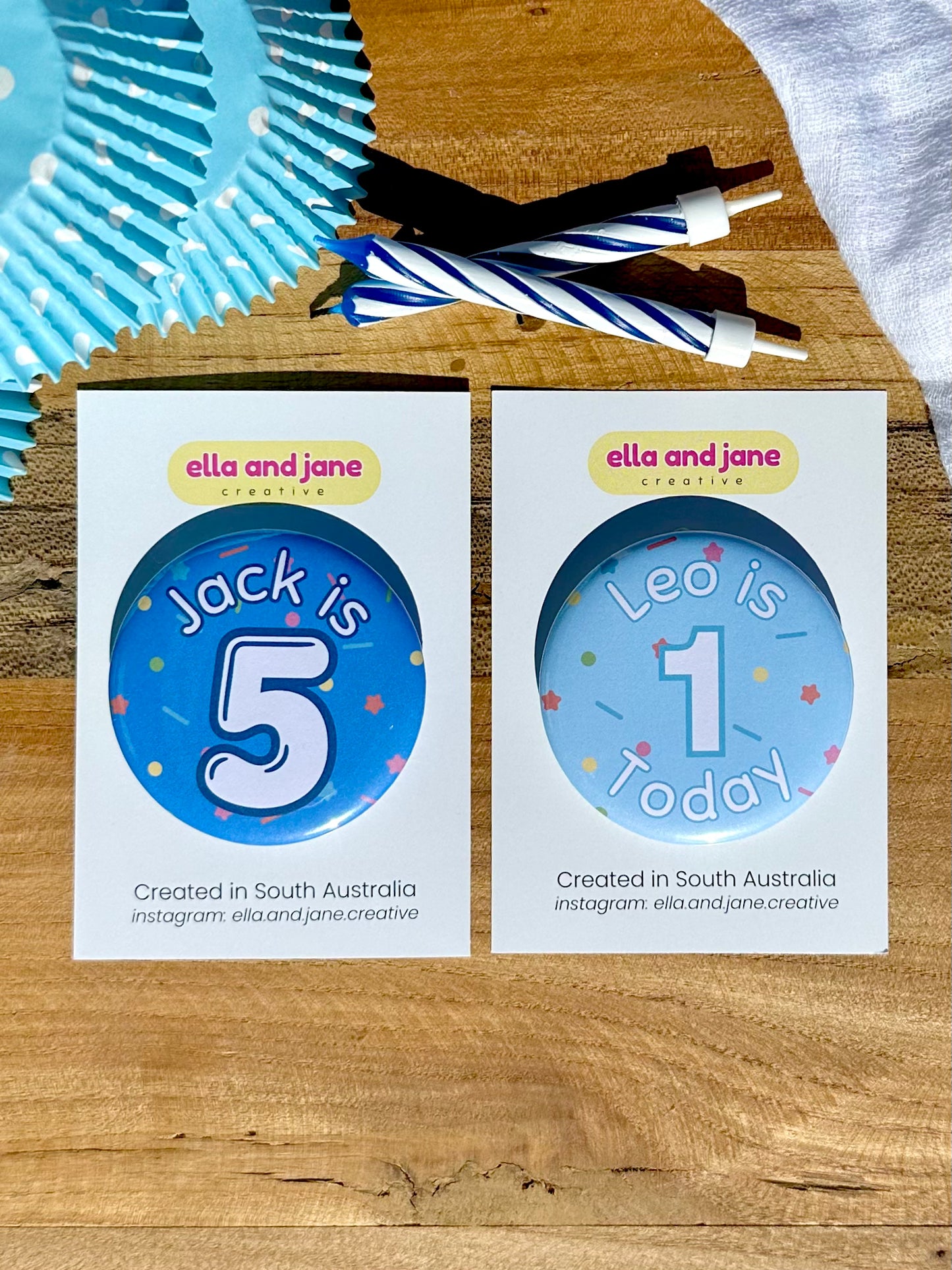 Blue birthday badges with sprinkles as the background. One is medium blue with the text 'Jack is 5' The other is light blue with the text 'Leo is 1 Today'. The badges are round and are sitting on a white product card. There are birthday party supplies as decorations, including cupcake cases and birthday candles.