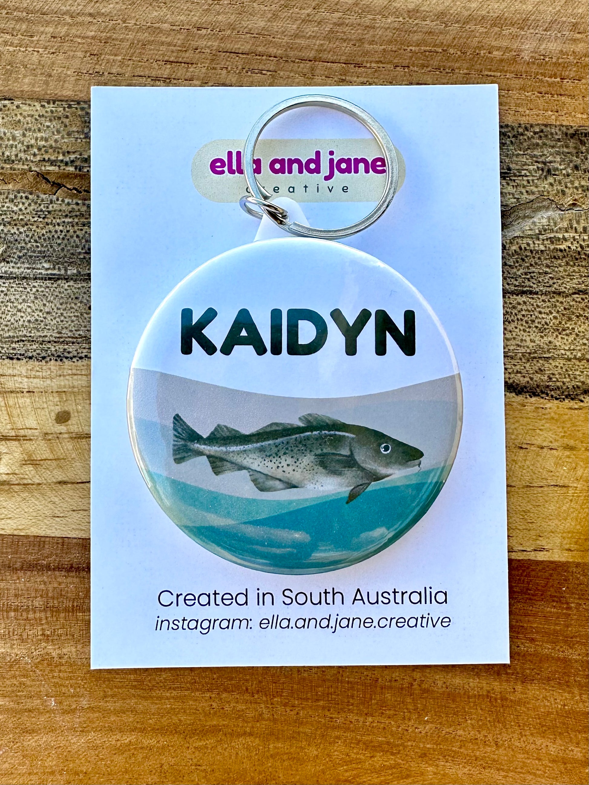 A round keyring with a white, grey and green wavy background. There is a name "Kaidyn' and a image of a murray cod fish. The keyring has a metal keyring attachment.