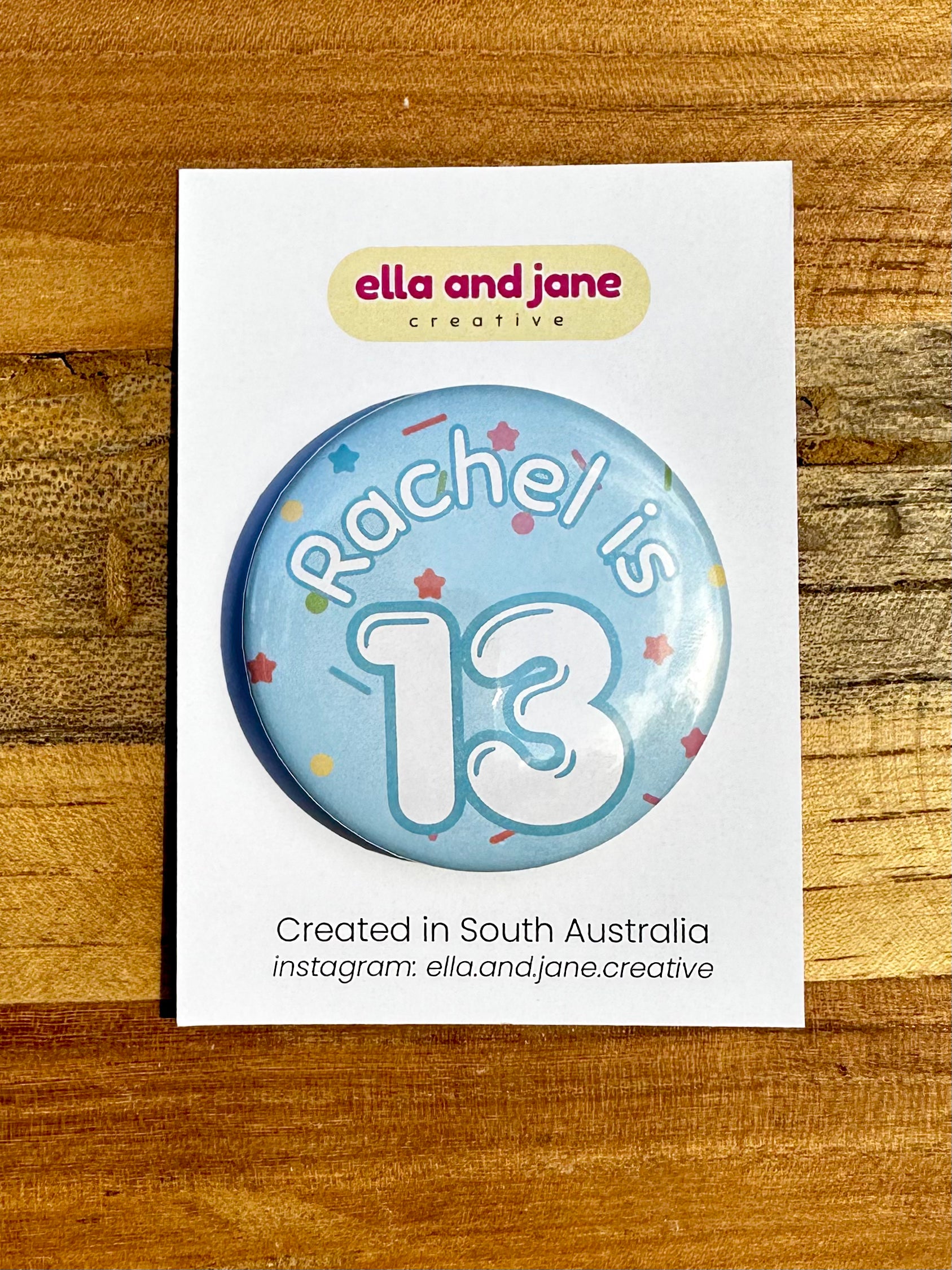 A light blue birthday badge with sprinkles as the background. The text says 'Rachel is 13' in white and blue. The badge is round and is sitting on a white product card. There are birthday party supplies as decorations, including cupcake cases and birthday candles.