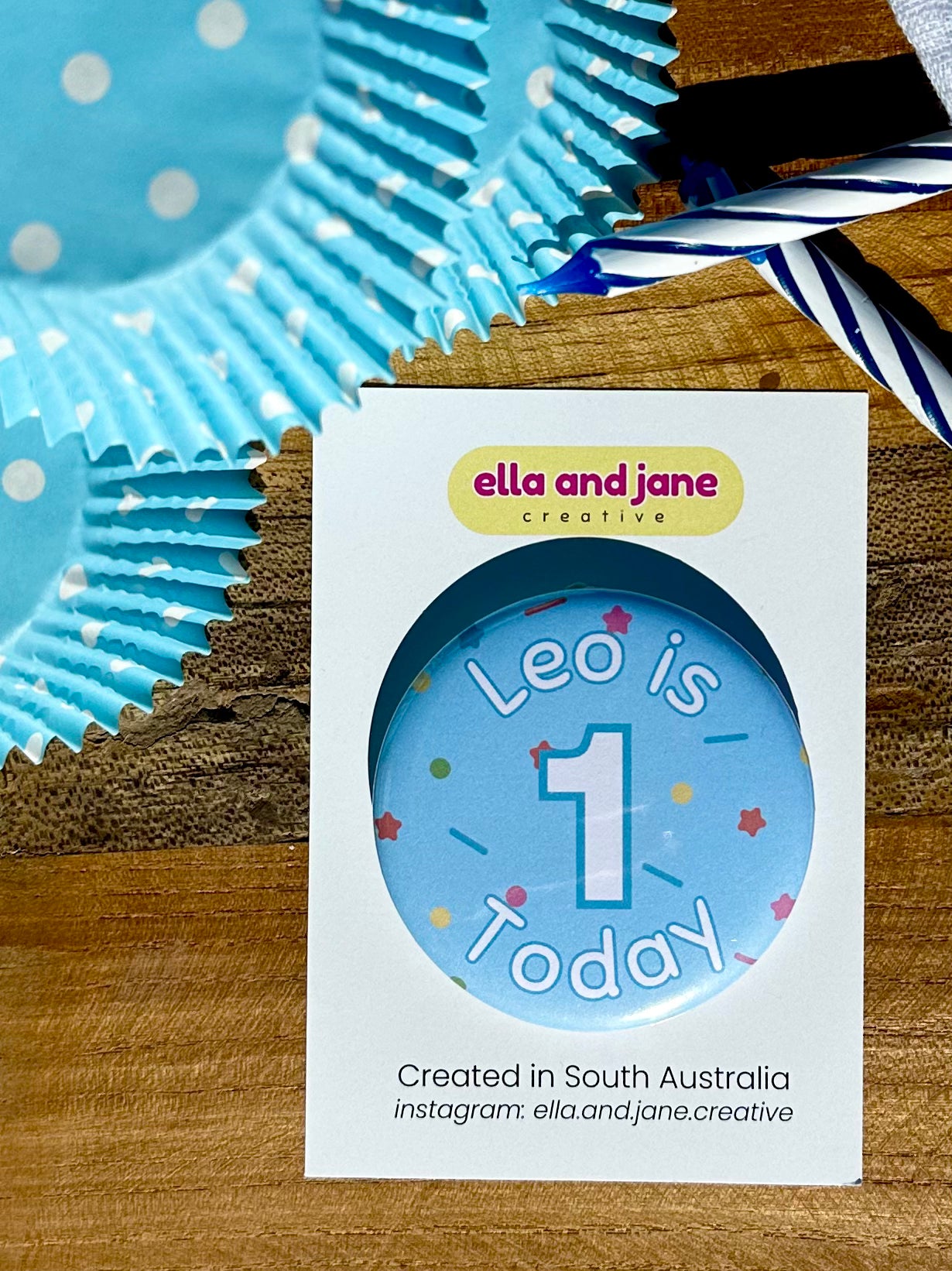 A light blue birthday badge with sprinkles as the background. The text says 'Leo is 1 Today' in white and blue. The badge is round and is sitting on a white product card. There are birthday party supplies as decorations, including cupcake cases and birthday candles.