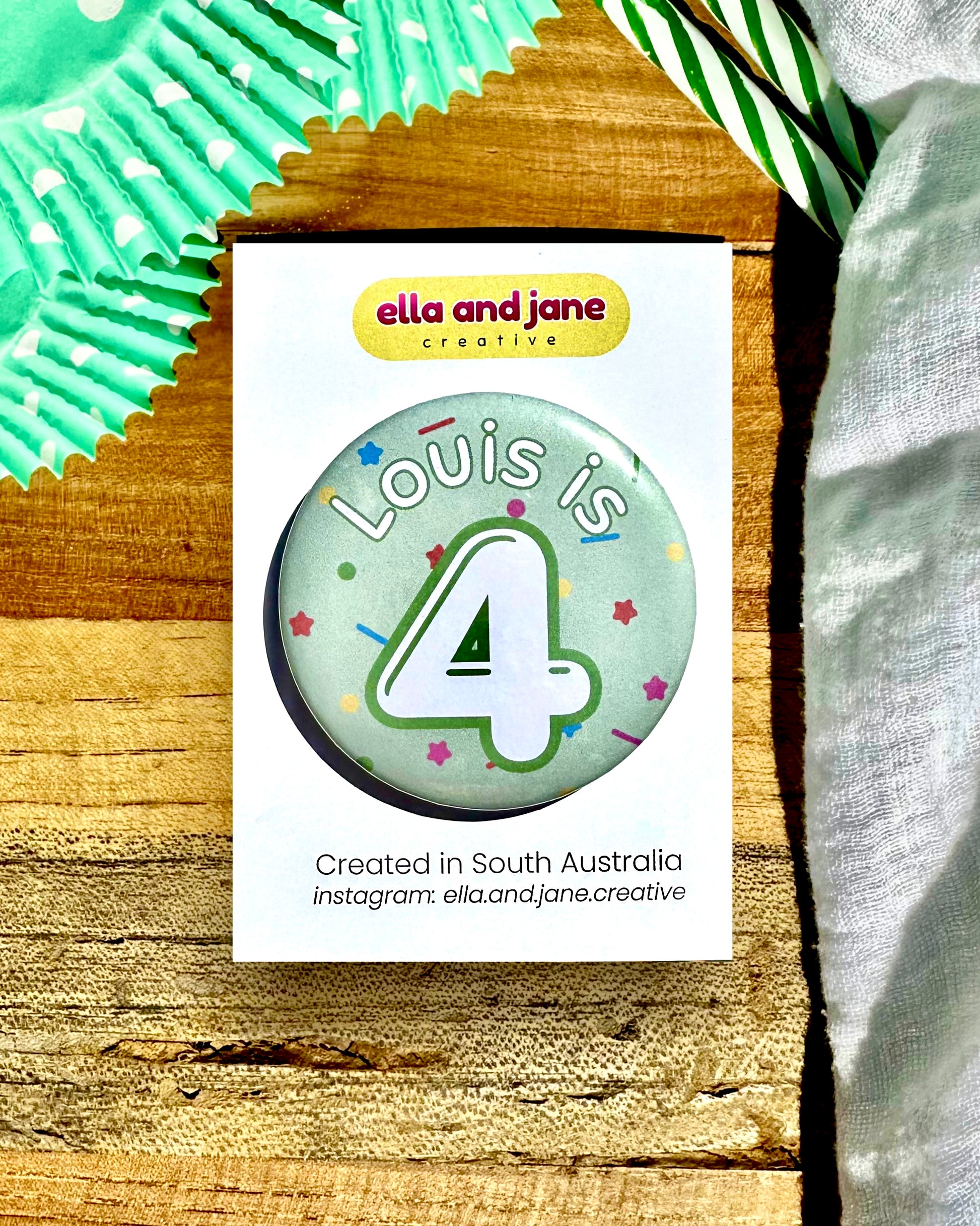 A pale green birthday badge with sprinkles as the background. The text says 'Louis is 4' in white and green. The badge is round and is sitting on a white product card. There are birthday party supplies as decorations, including cupcake cases and birthday candles.