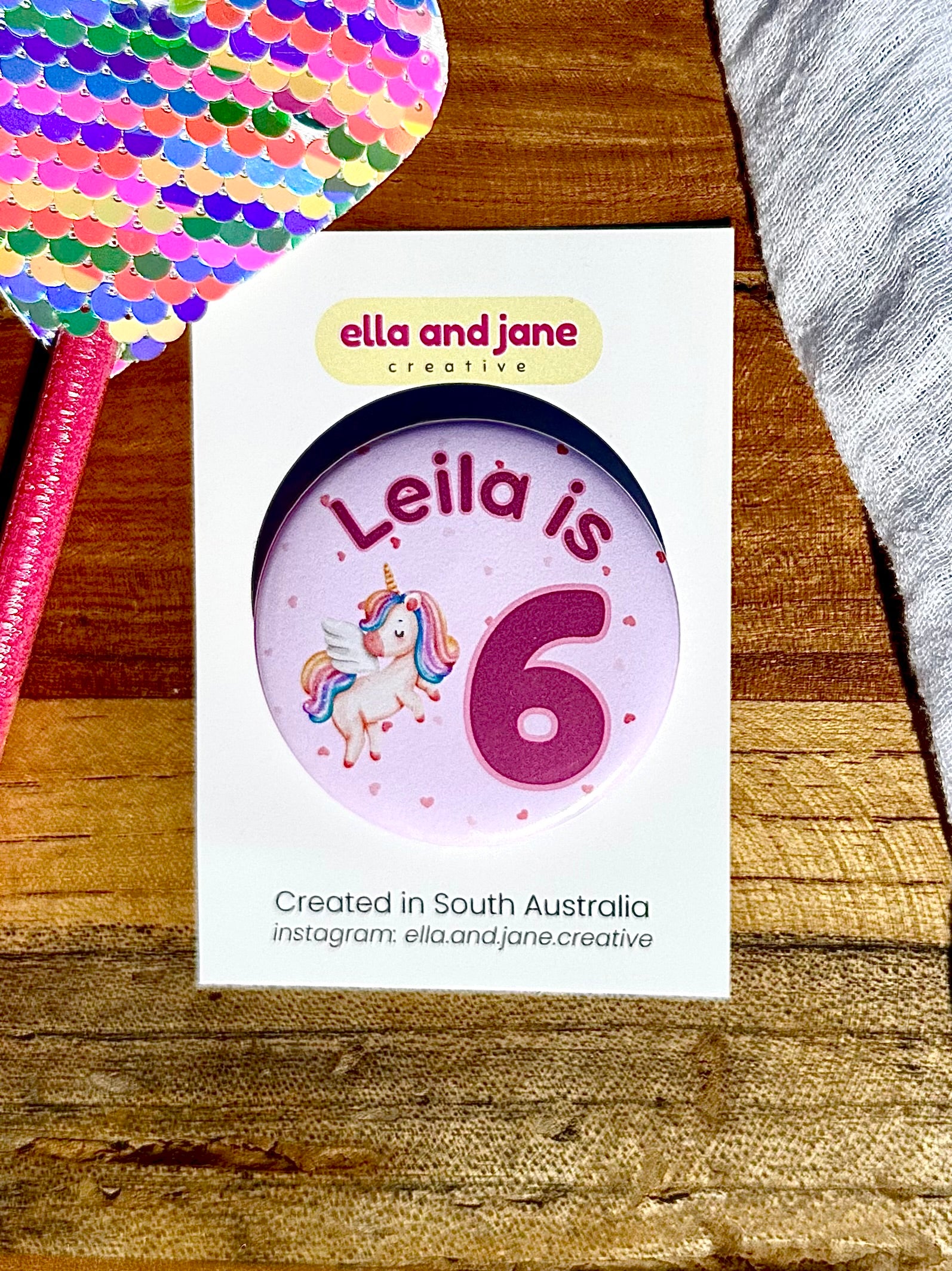 A unicorn party badge which is light pink with darker pink hearts as the background. There is a flying unicorn with pastel rainbow tail and mane. The text is Leila is 6. The badge is on a white product card and laying on a piece of wood. There are rainbow decorations on the wood.