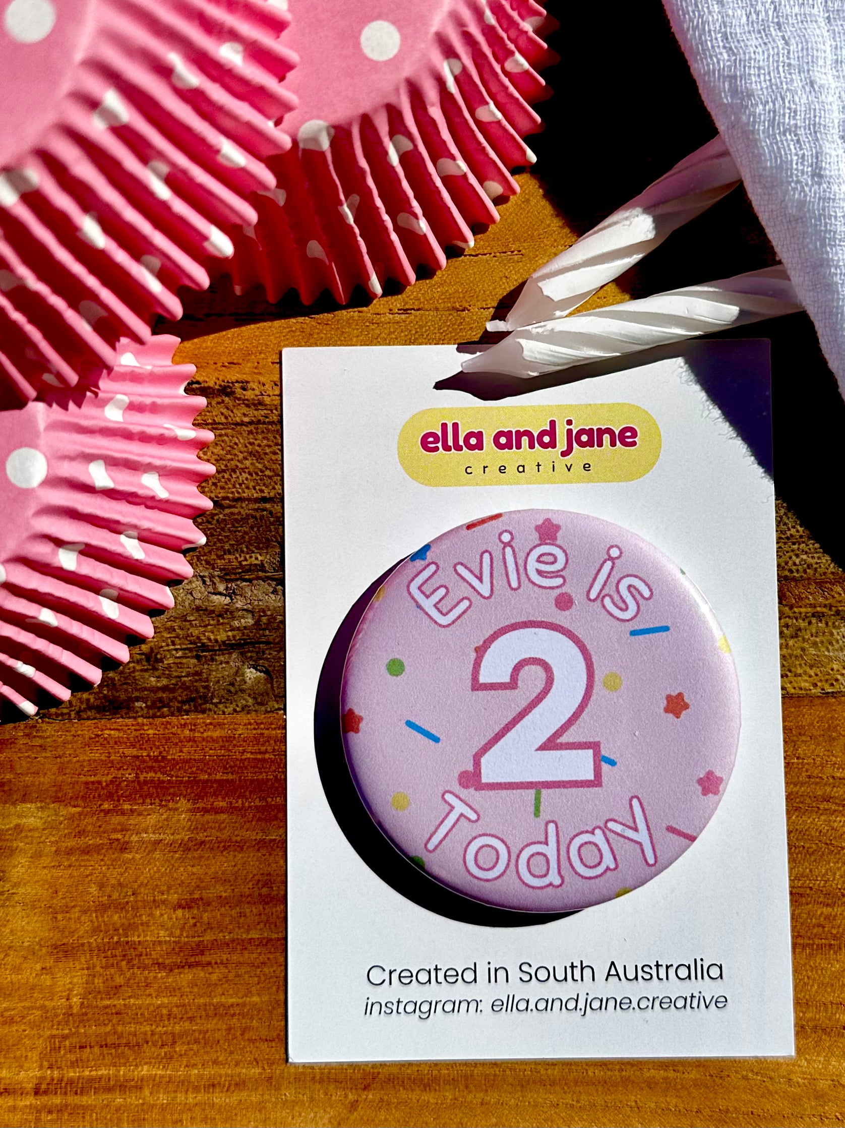 A light pink birthday badge with sprinkles as the background. The text says 'Evie is 2 Today' in white and pink. The badge is round and is sitting on a white product card. There are birthday party supplies as decorations, including cupcake cases and birthday candles.