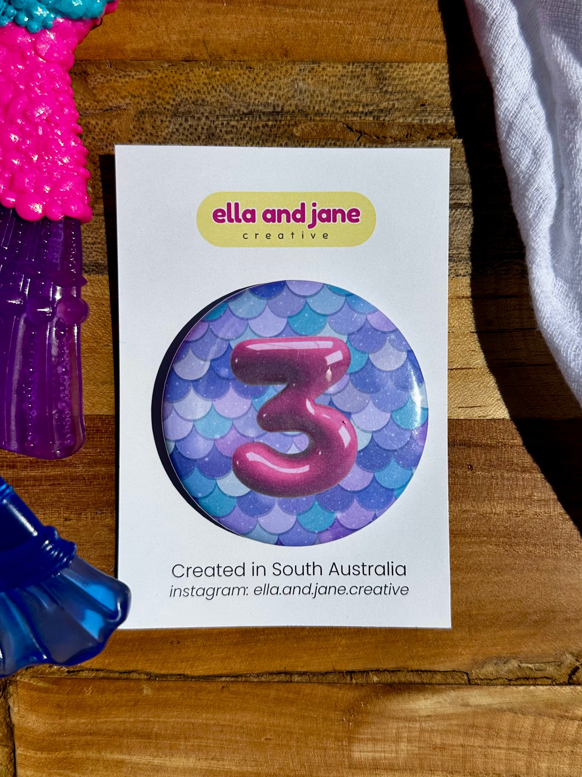 A blue and green mermaid tail birthday badge. It has the number 3 in pink written on it and the party badge is sitting on a product card and laying on a piece of wood with decorations.