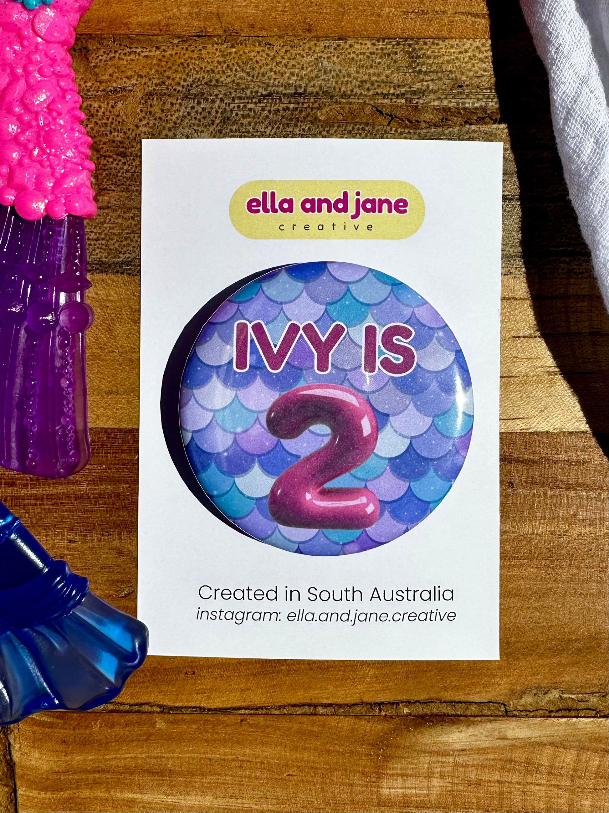 A blue and green mermaid tail birthday badge. It has custom text on the badge 'Ivy is 2', the party badge is sitting on a product card and laying on a piece of wood with decorations.