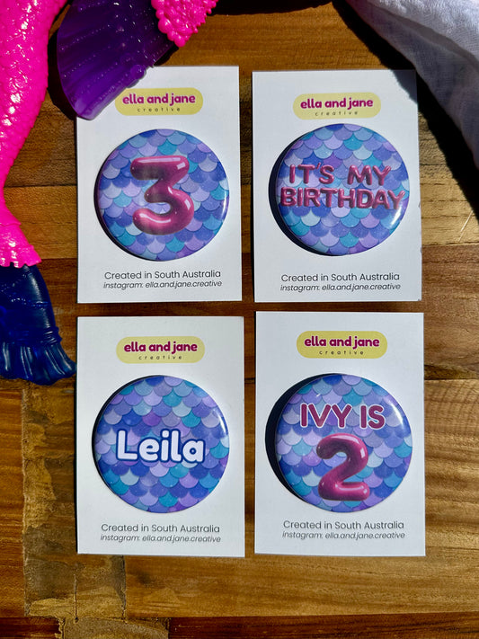 Four purple, blue and green mermaid tail birthday badges. The first badge has the number 3 for a 3rd birthday, The second has the text It's My Birthday, the third has the name 'Leila' for a name badge and the fourth is personalised with Ivy is 2. 
The party badges are sitting on a product card and laying on a piece of wood with decorations.