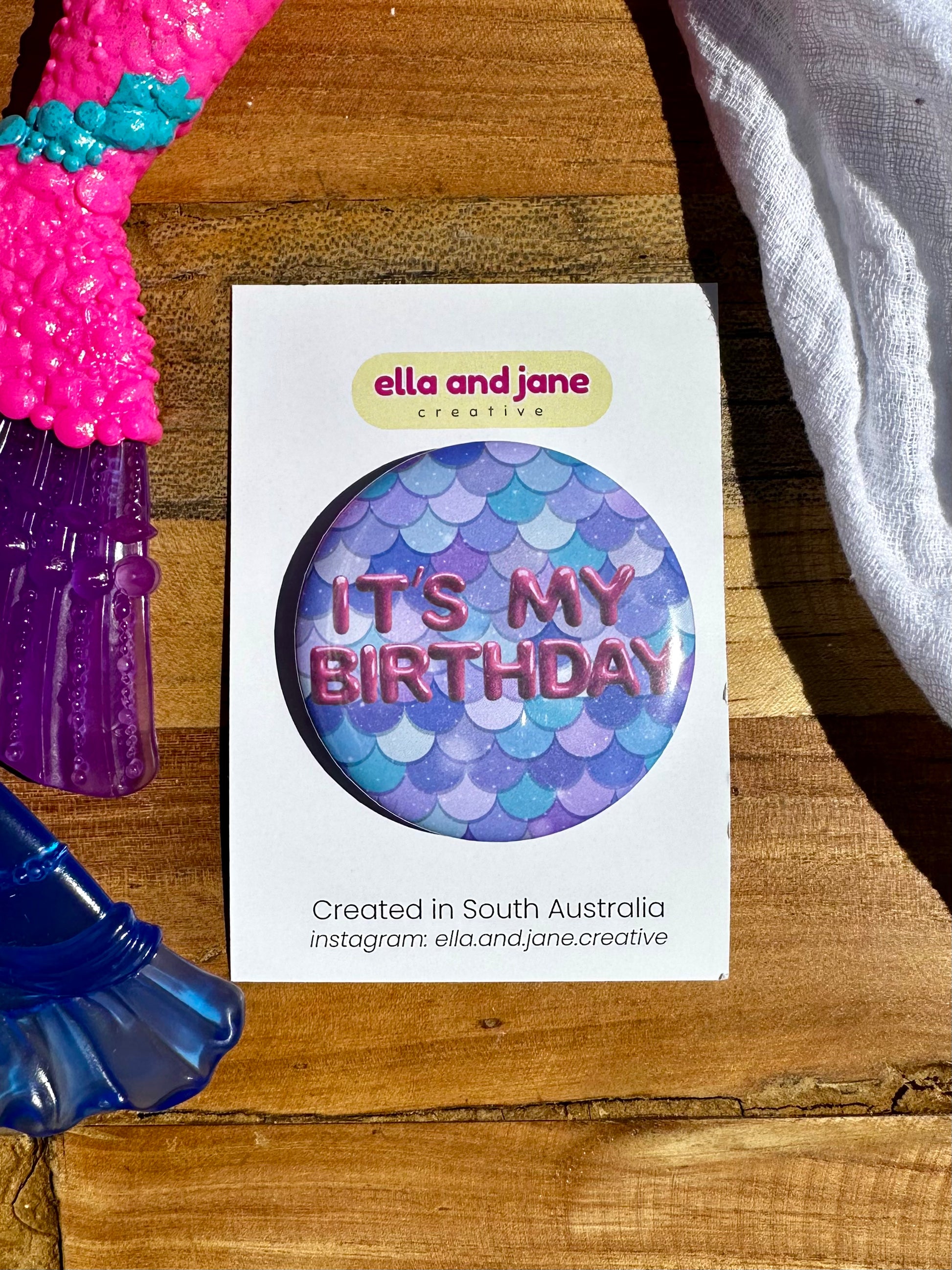 A blue and green mermaid tail birthday badge. It has the text It's My Birthday, the party badge is sitting on a product card and laying on a piece of wood with decorations.
