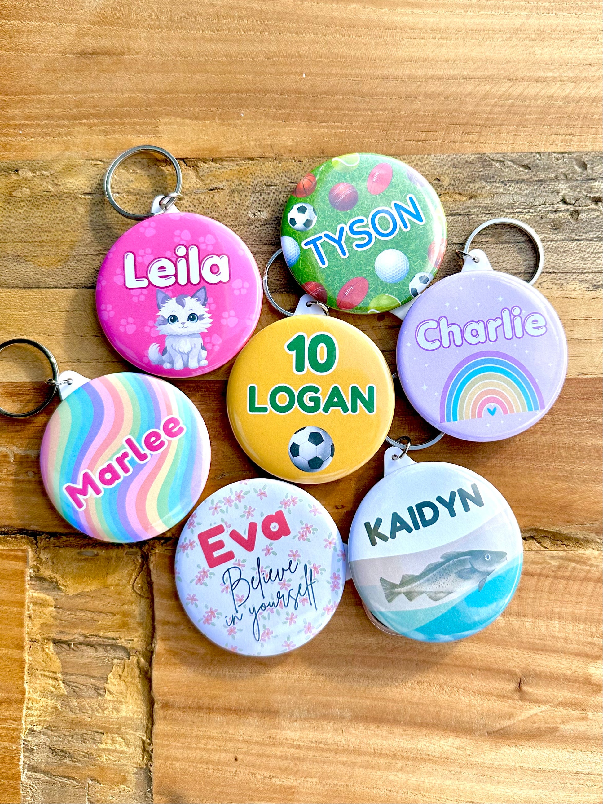 7 personalised keyrings with a name and image. One has a pink background with paw prints and a white and grey cat. One has green lawn and various sporting balls. One has a light purple background with a colourful rainbow. One has white grey and grey waves with a murray cod fish. One has a white background with soft pink flowers. One has a rainbow swirl background.