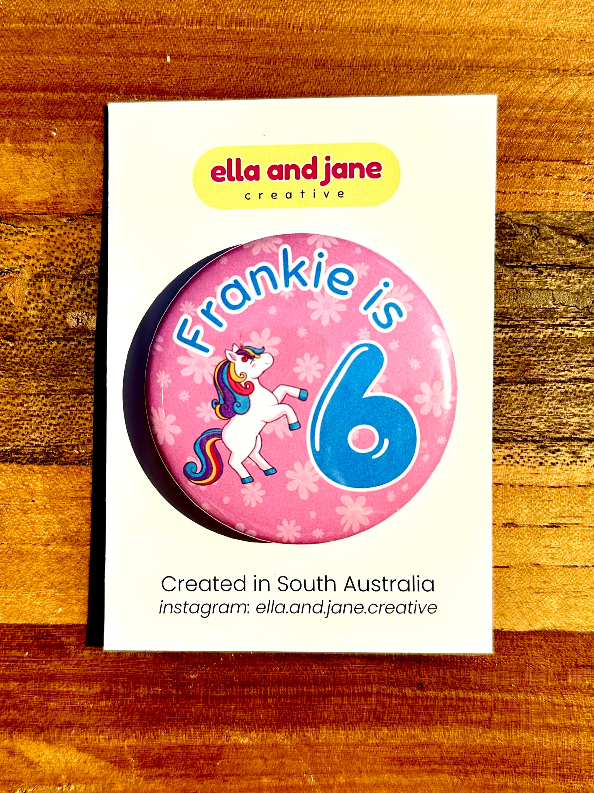 A round party badge which is medium pink with lighter pink flowers as the background. There is a unicorn with darker rainbow tail and mane. The text is in blue and says Frankie is 6. The badge is on a white product card and laying on a piece of wood.