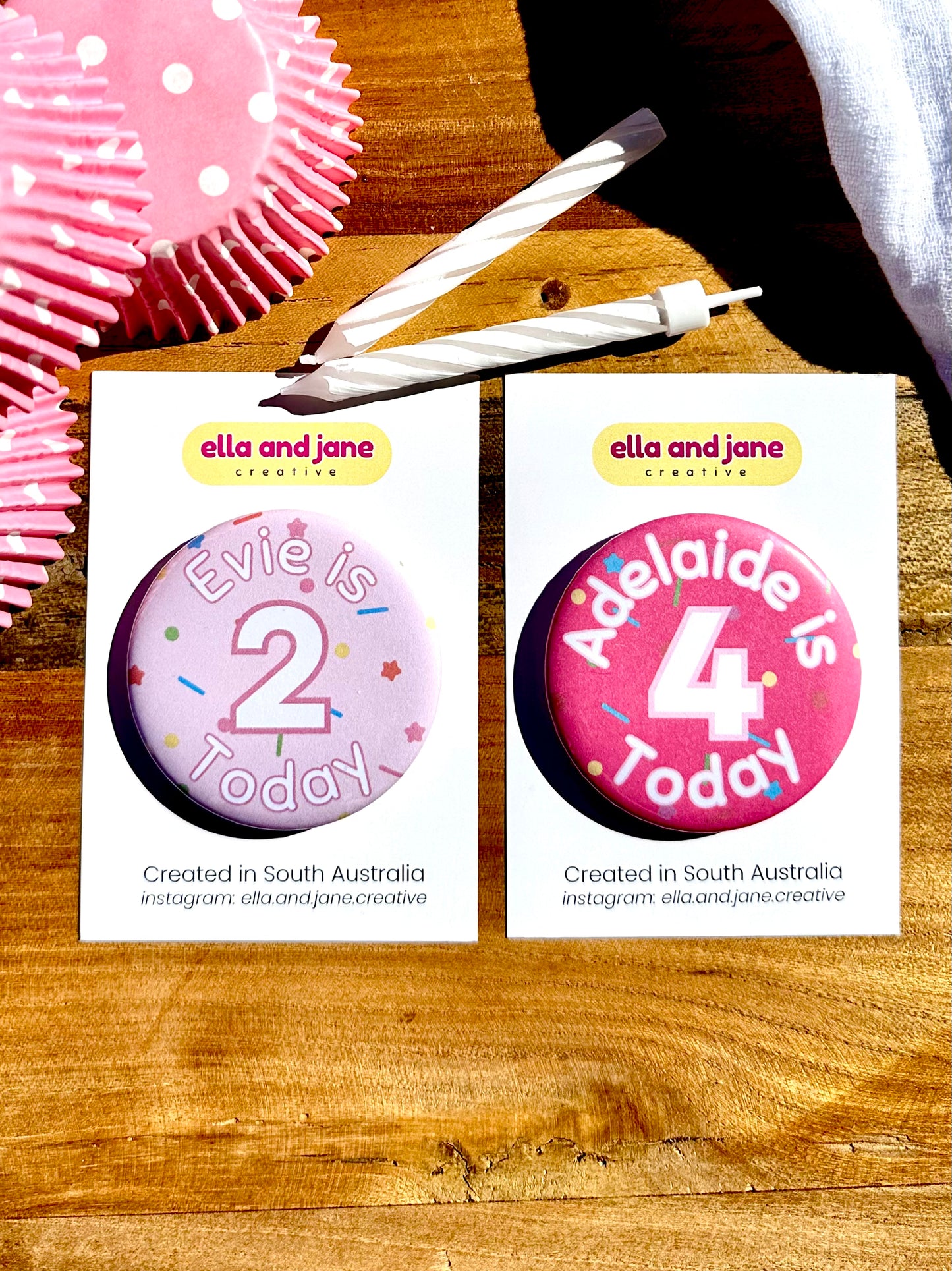 Pink birthday badges with sprinkles as the background. One is light pink with the text 'Evie is 2 Today' and the other is medium pink with the text 'Adelaide is 4 Today' The badges are round and are sitting on a white product card. There are birthday party supplies as decorations, including cupcake cases and birthday candles.