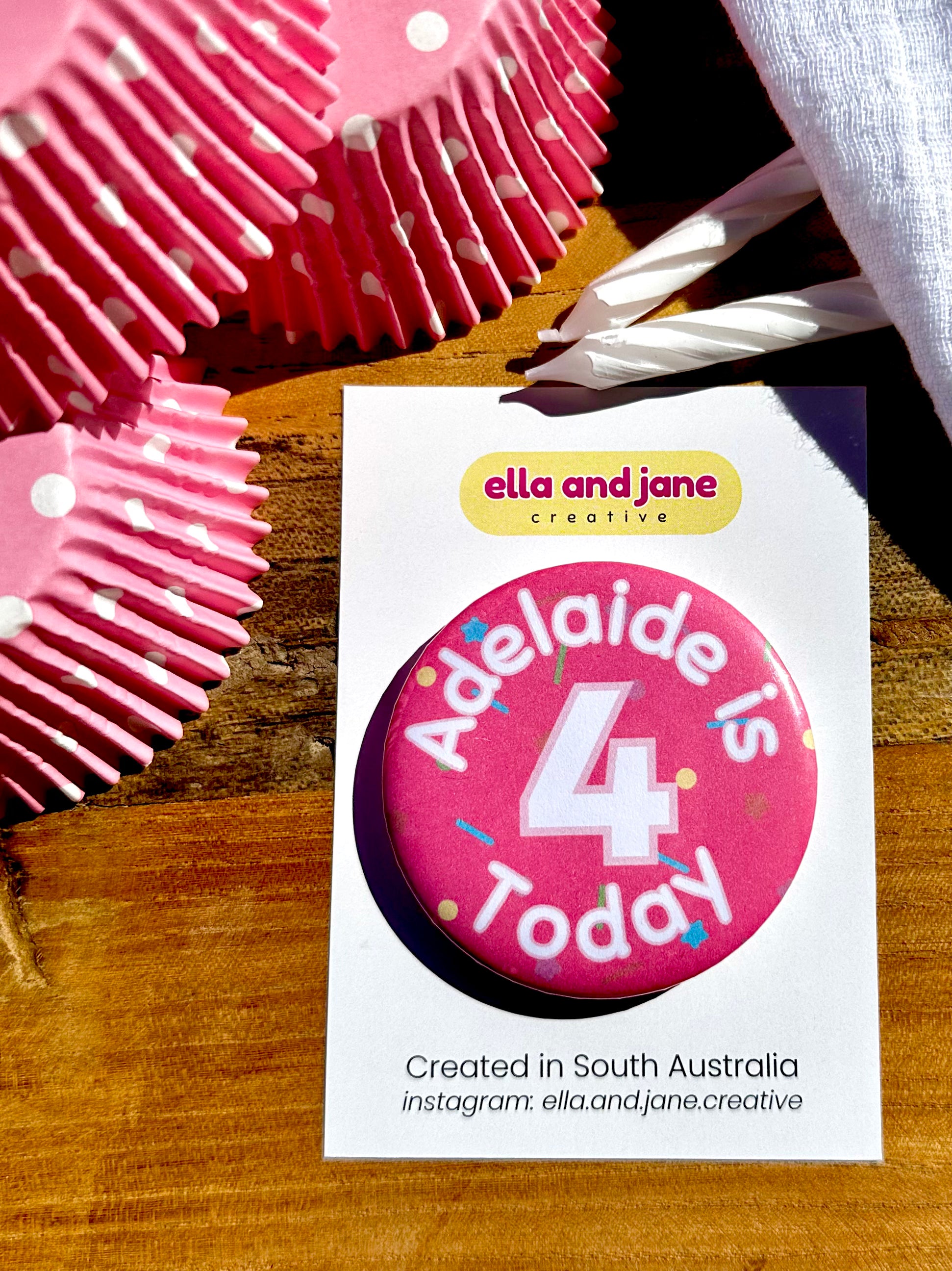 A medium pink birthday badge with sprinkles as the background. The text says 'Adelaide is 4 Today' in white and pink. The badge is round and is sitting on a white product card. There are birthday party supplies as decorations, including cupcake cases and birthday candles.