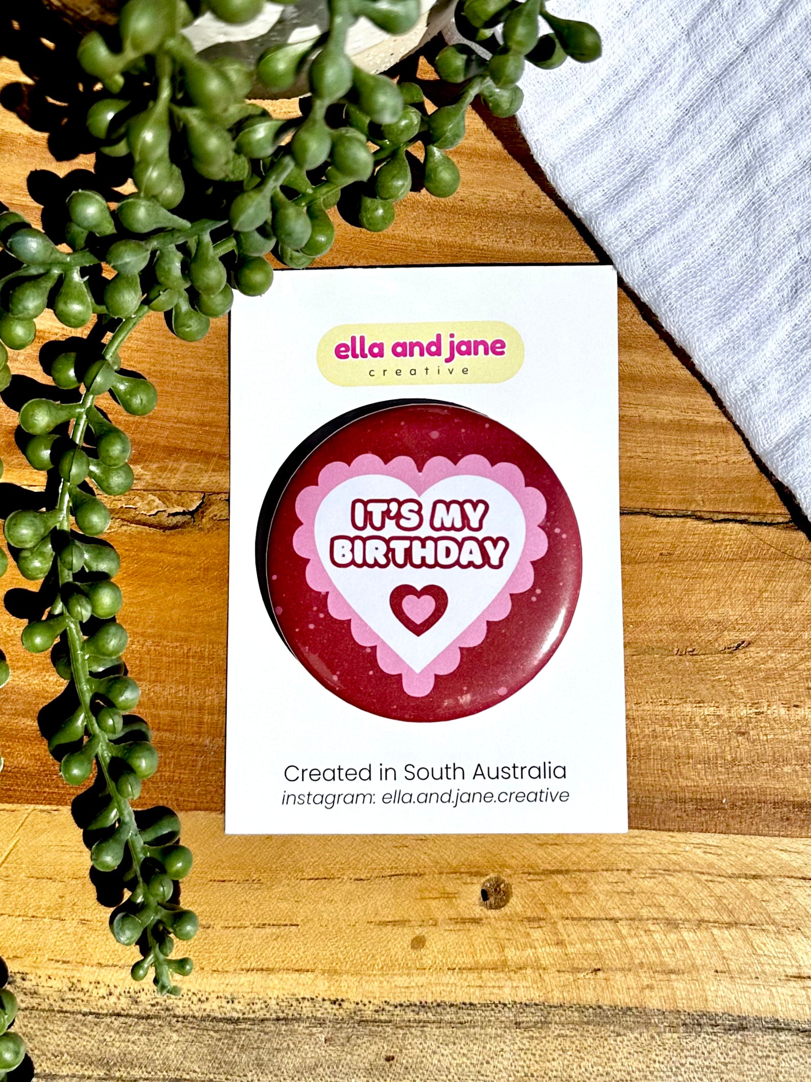 Pink and red hearts birthday badges. The party badge is a deep red maroon colour with light pink spots. There is a white heart in the centre with pink bubble ripples around the edge of the heart. There is text 'It's My Birthday' in the centre of the white heart. There is another smaller heart under the text which is burgundy and pink. The birthday badge is sitting on white product cards and laying on a piece of wood with decorations.