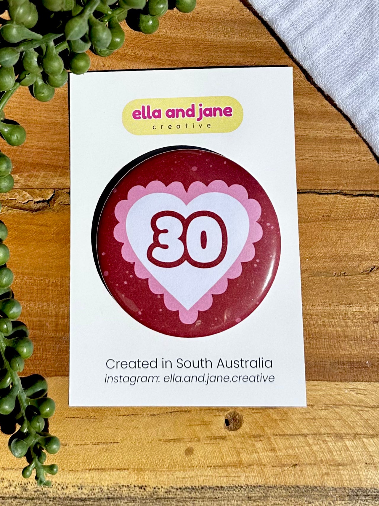 Pink and red hearts birthday badges. The party badge is a deep red maroon colour with light pink spots. There is a white heart in the centre with pink bubble ripples around the edge of the heart. There is text '30' in the centre of the white heart.  The birthday badge is sitting on white product cards and laying on a piece of wood with decorations.