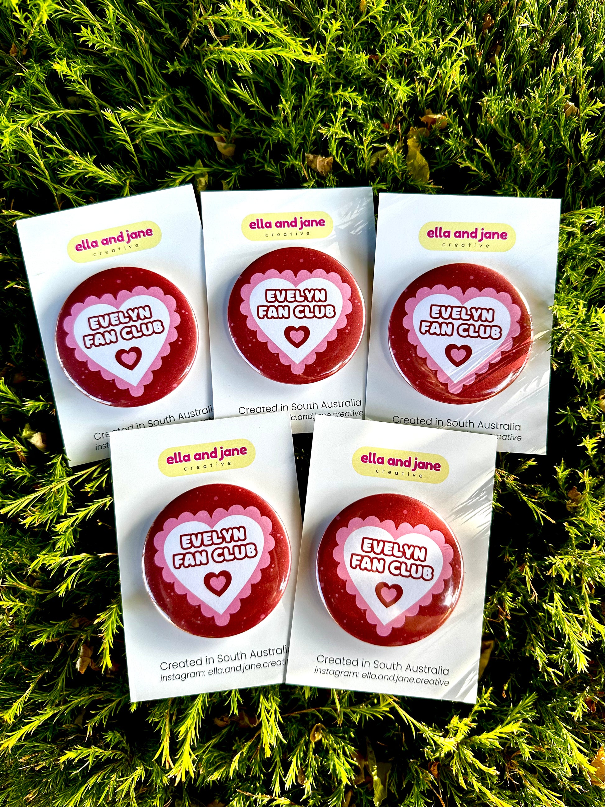 Pink and red hearts birthday badges. The party badges are a deep red maroon colour with light pink spots. There is a white heart in the centre with pink bubble ripples around the edge of the heart. There is text 'Evelyn Fan Club' in the centre of the white heart. There is an additional heart under the text with maroon and pink in the centre. The birthday badge is sitting on white product cards and laying on a piece of wood with decorations. The five badges are for party favours.