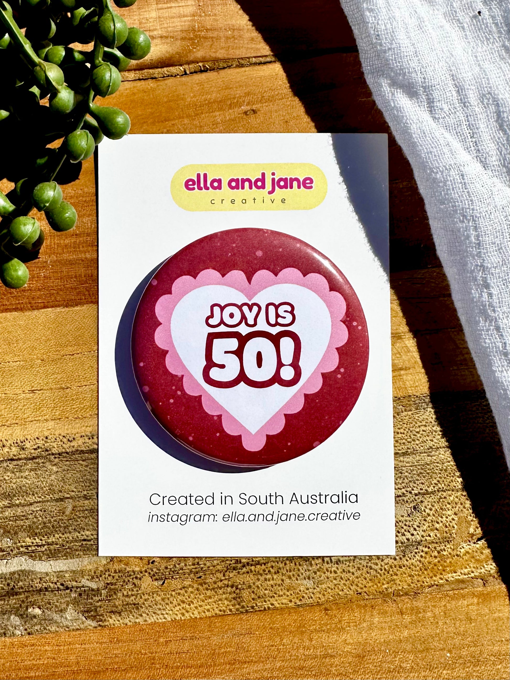 Pink and red hearts birthday badge. The party badge is a deep red maroon colour with light pink spots. There is a white heart in the centre with pink bubble ripples around the edge of the heart. There is text 'Joy is 50!' in the centre of the white heart.  The birthday badge is sitting on white product cards and laying on a piece of wood with decorations.