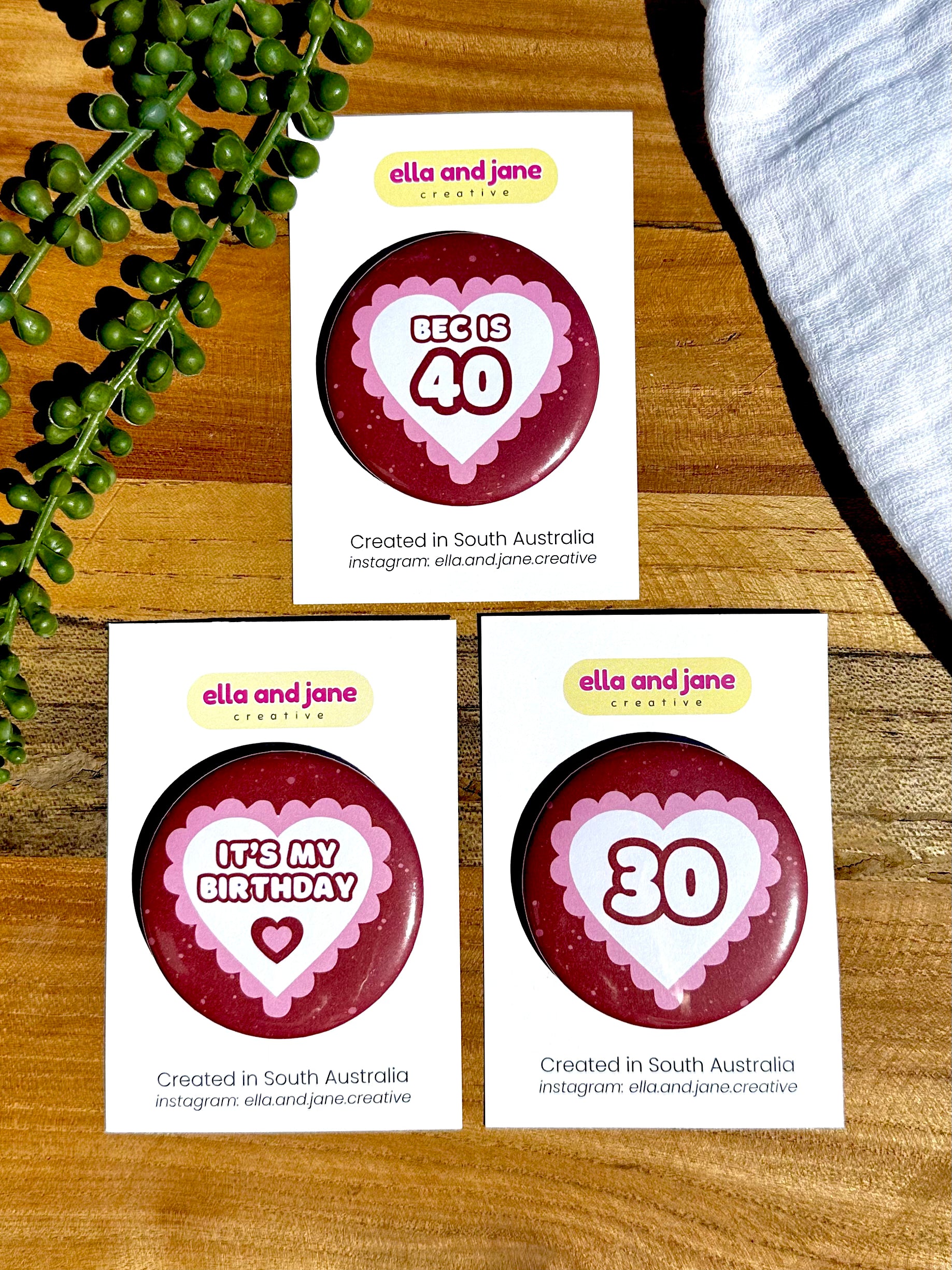 Three pink and read hearts birthday badges. The party badges are a deep red maroon colour with light pink spots. There is a white heart in the centre with pink bubble ripples around the edge of the heart. There is text in the middle. The first badge says 'Bec is 40' the second badge says 'It's my birthday' with an additional smaller heart underneath of maroon and pink. The third badge has the text '30'. The birthday badges are sitting on white product cards and laying on a piece of wood with decorations.