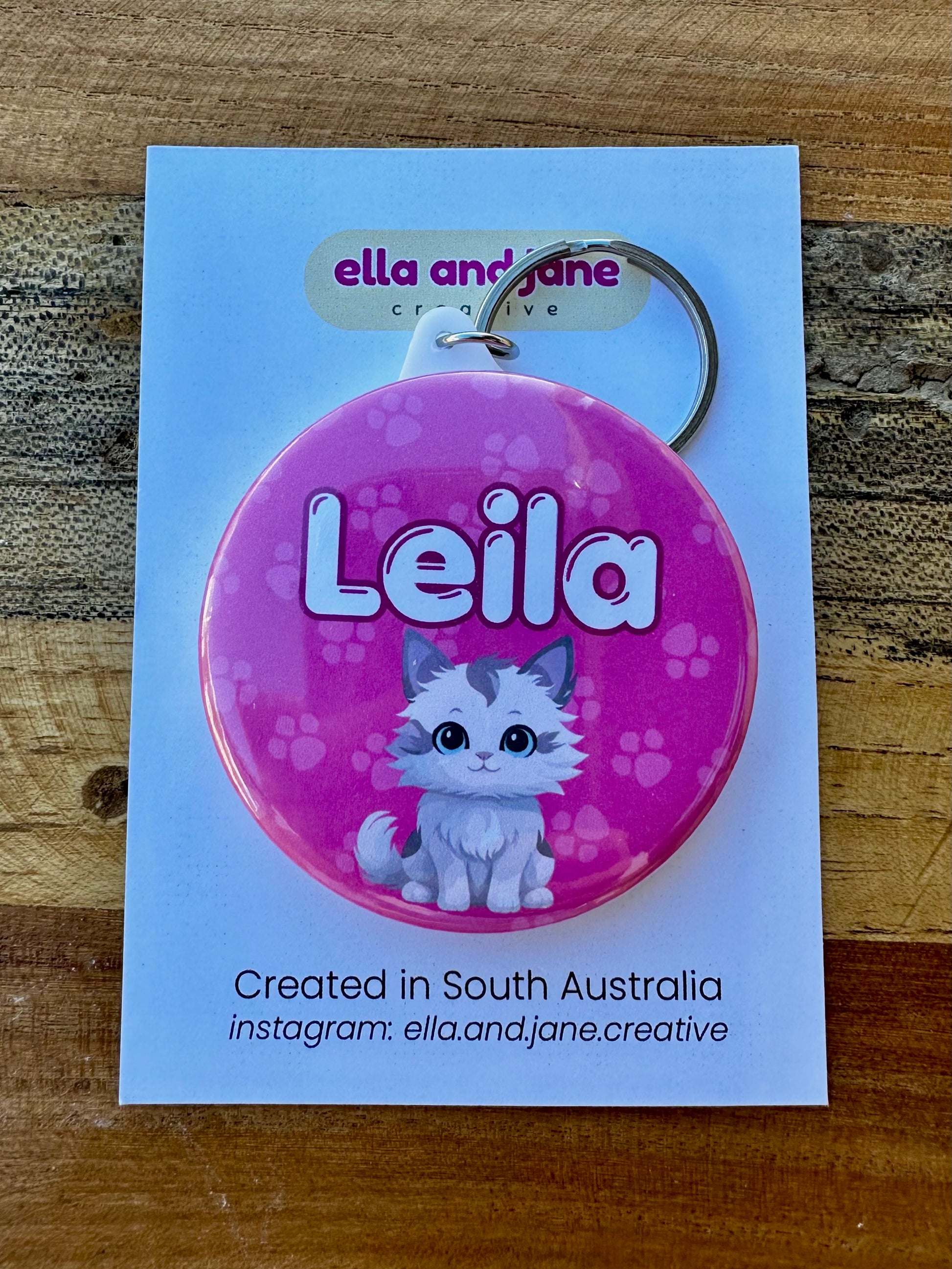 A round keyring with a medium pink background and light pink paw prints. There is a name 'Leila' in white with a dark pink border and an image of a white and grey fluffy cat. The keyring has a metal keyring attachment, and it is laying on a white product card on top of a wooden block.