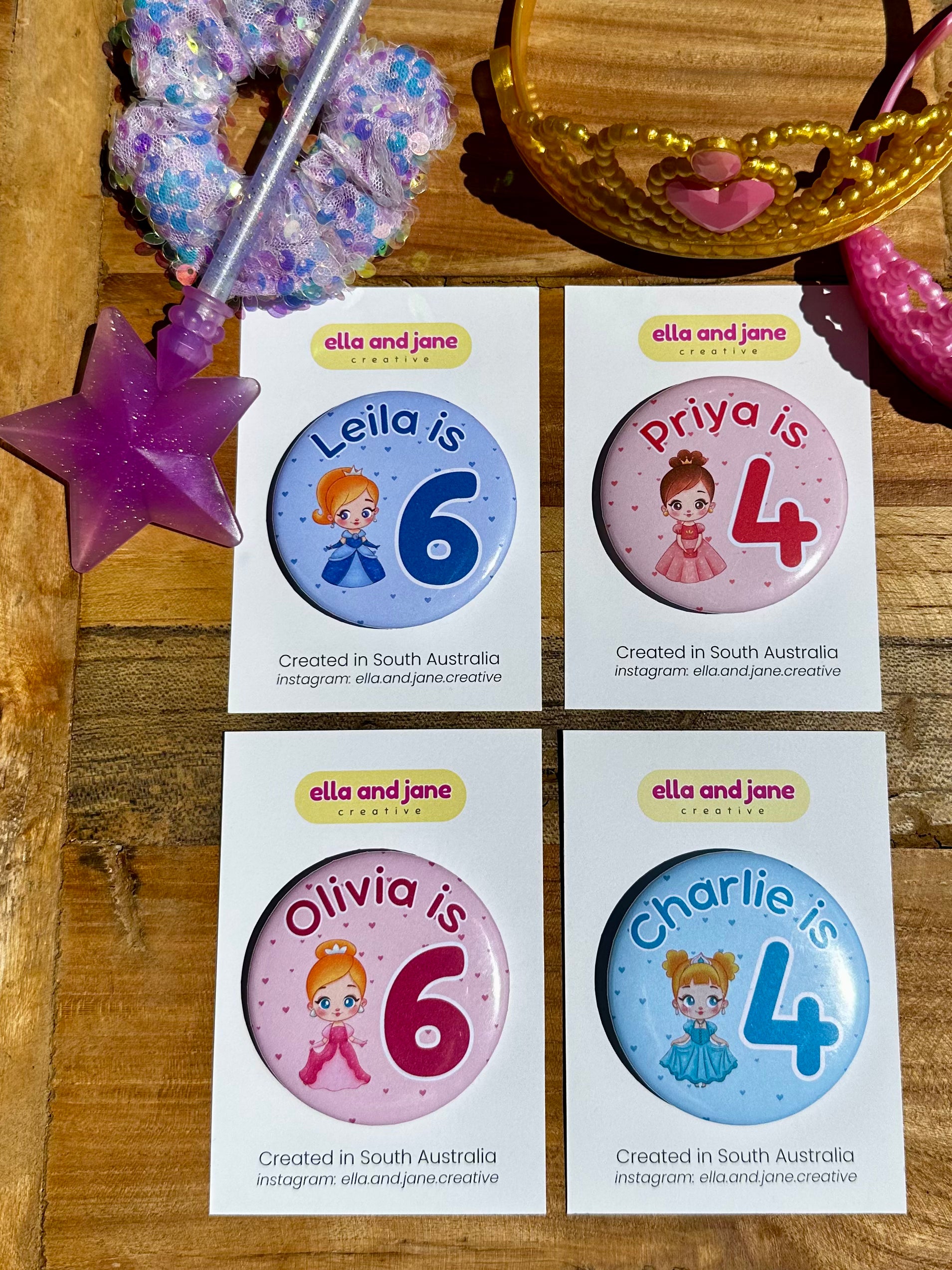 Four custom birthday badges. The first is blue with the text 'Leila is 6' and a princess with a blue dress. The second is a light pink birthday badge with the text 'Priya is 4' and a princess with a light pink orange dress. The third badge is pink with the text 'Olivia is 6' and a princess wearing a pink dress. The fourth badge is blue with the text 'Charlie is 4' and a princess wearing a crown and a light blue dress. The party badges are decorated with princess crowns and wands on a piece of wood.