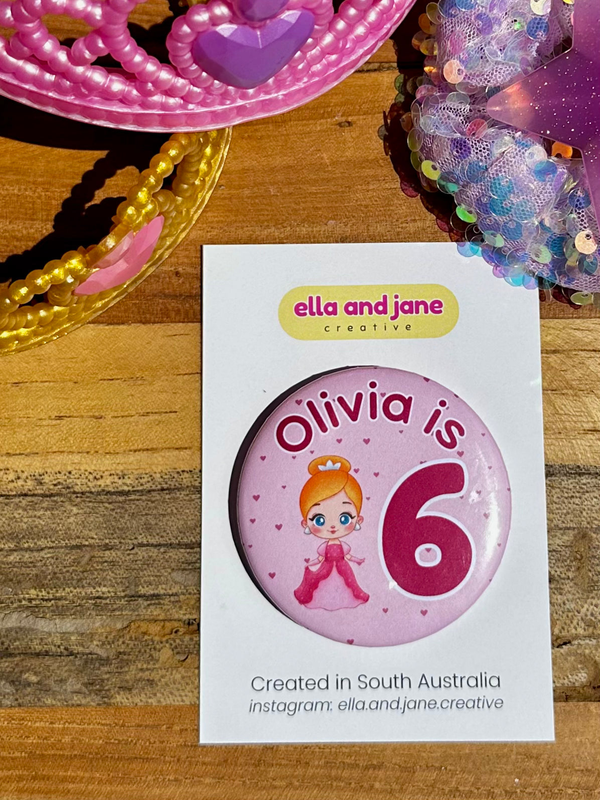 Custom birthday party badge with the text 'Olivia is 6' with the 6 being dark pink and large. There is also a picture of a princess wearing a pink dress and a crown. There are love hearts in the background. The party badges are decorated with princess crowns and wands on a piece of wood.