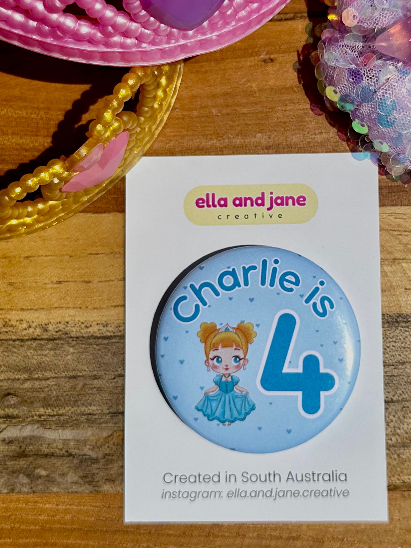 Blue custom birthday party badge with the text 'Charlie is 4' with the 4 being Blue and large. There is also a picture of a princess wearing a blue dress and a crown. There are love hearts in the background. The party badges are decorated with princess crowns and wands on a piece of wood.