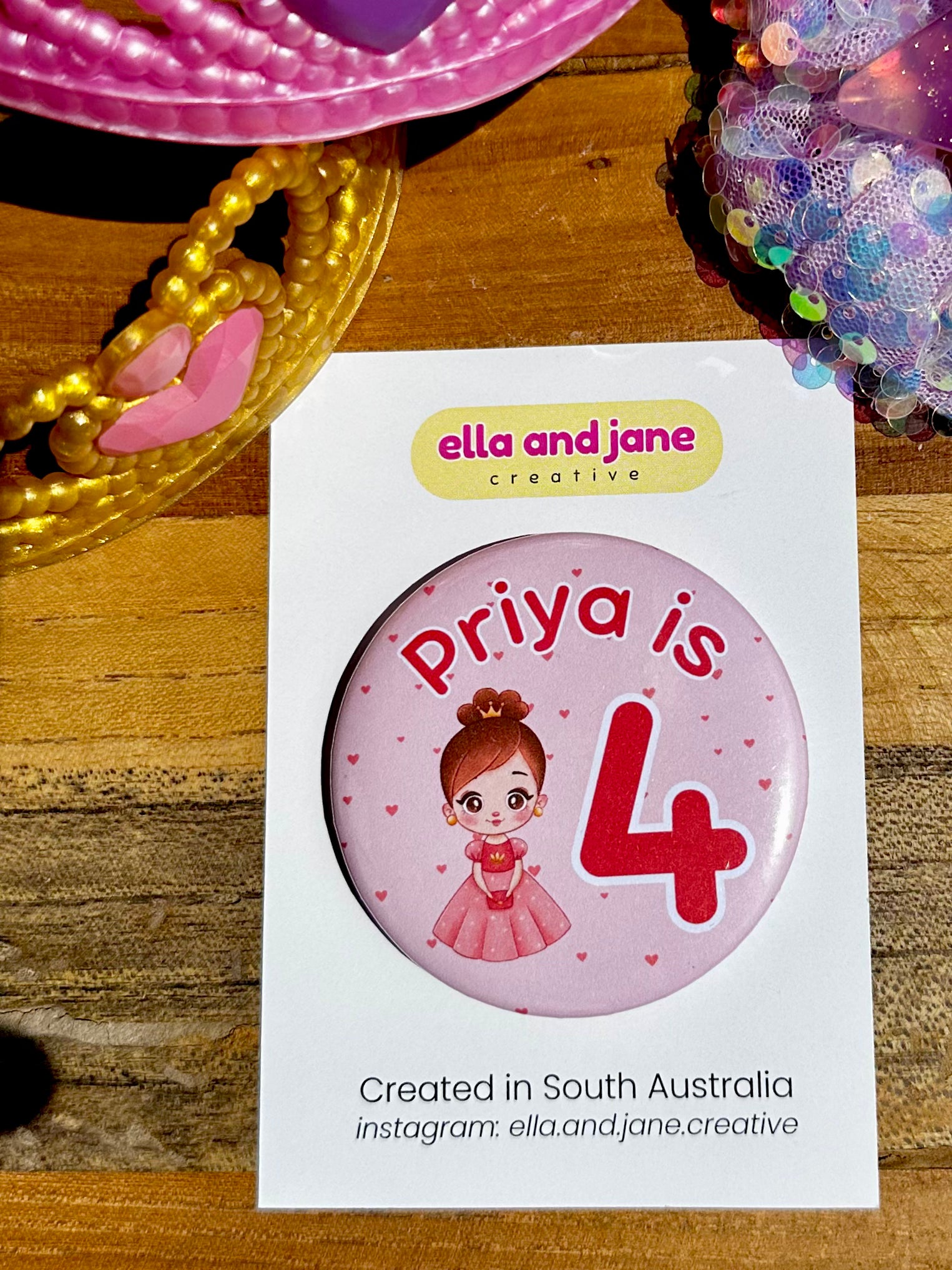 Custom birthday party badge with the text 'Priya is 4' with the 4 being Orange/Pink and large. There is also a picture of a princess wearing a pink and orange dress and a crown. There are love hearts in the background. The party badges are decorated with princess crowns and wands on a piece of wood.