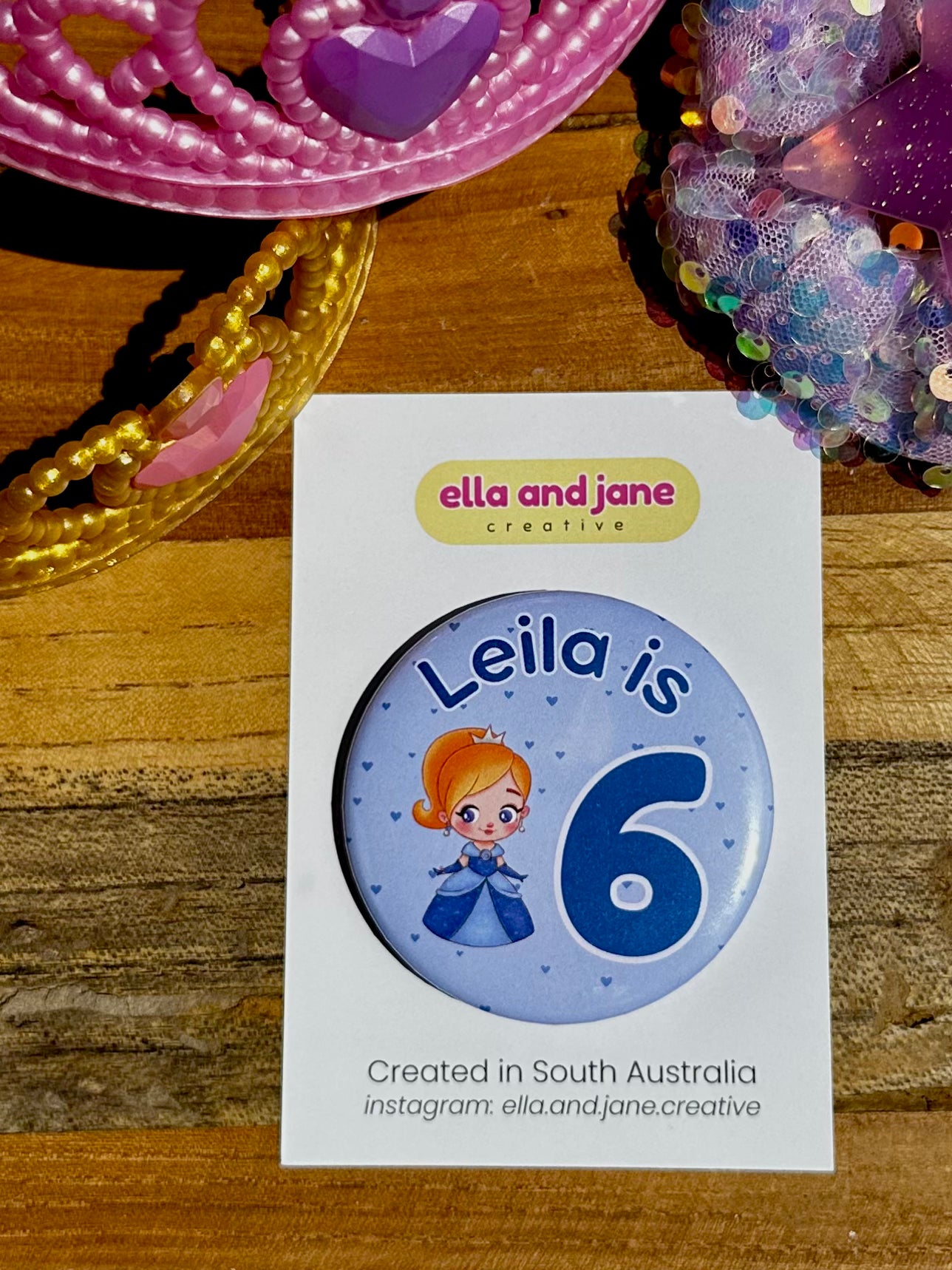 Blue custom birthday party badge with the text 'Leila is 6' with the 6 being dark blue and large. There is also a picture of a princess wearing a blue dress and a crown. There are love hearts in the background. The party badges are decorated with princess crowns and wands on a piece of wood.