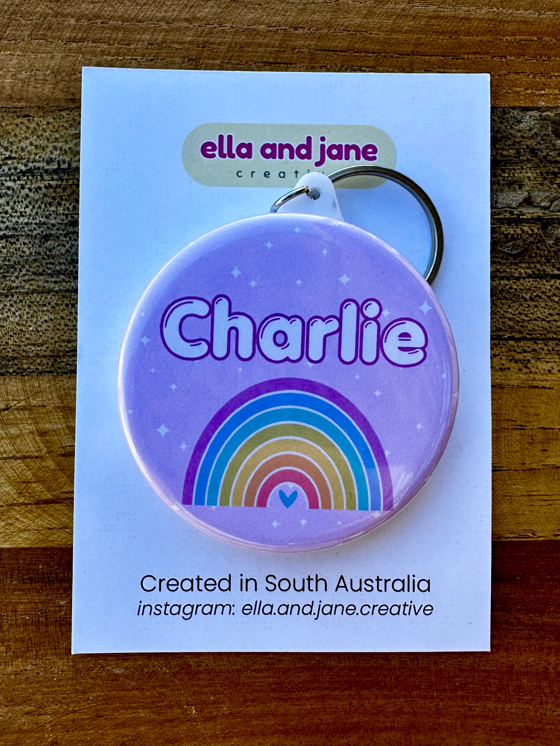 Custom keyring and bag tag with a light lilac purple background with white stars. There is a name 'Charlie' as well as a rainbow and a small love heart. The key ring is attached with a metal keyring hook and is sitting on a white product card which is laying on a wooden block.