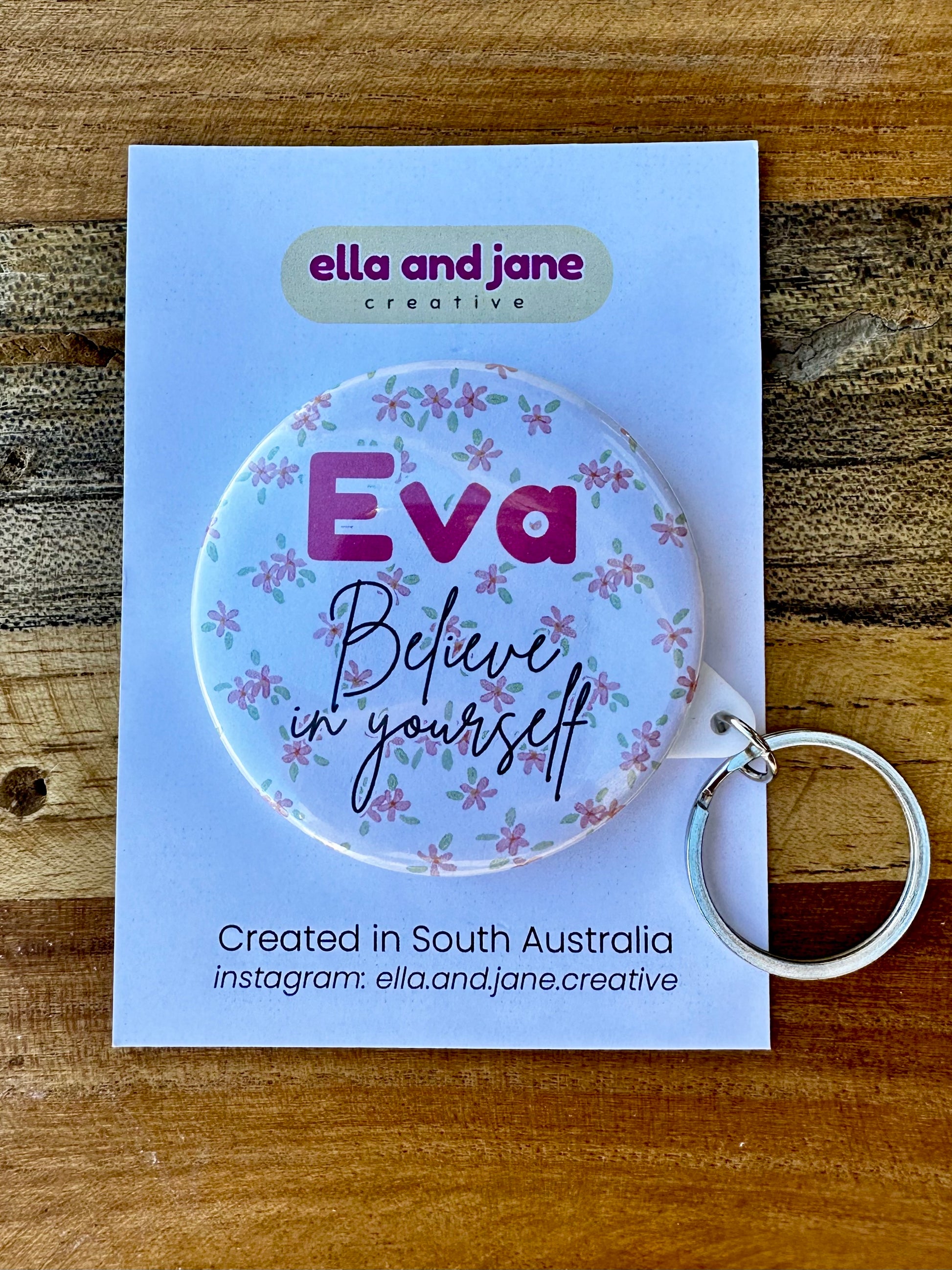 A round keyring with a white background and soft pink flowers. There is a name 'Eva' and the words 'Believe in yourself'. The keyring has a metal keyring attachment.