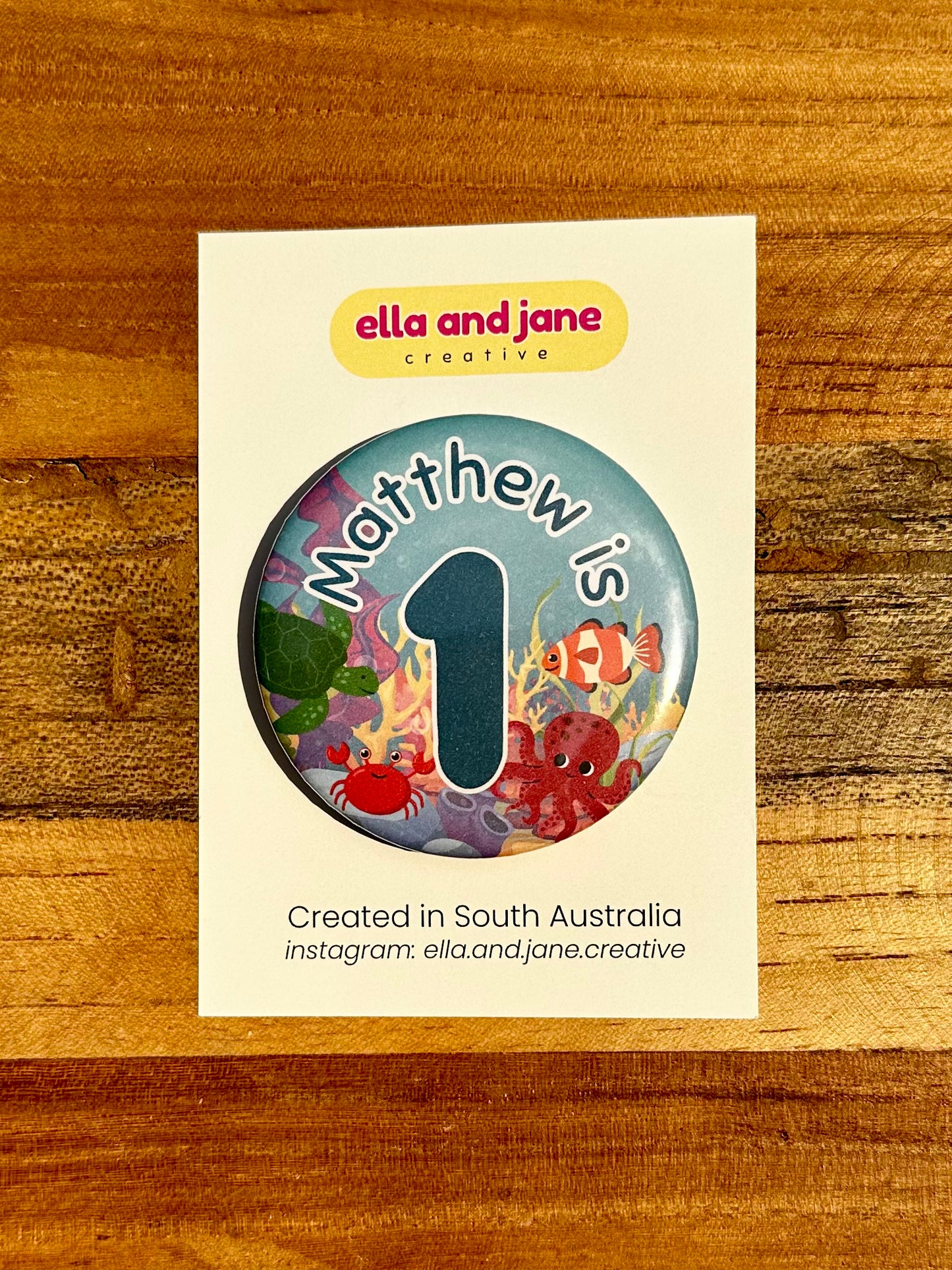 A round birthday party badge with the text Matthew is 1 in dark blue. The background image is under the sea with water, seaweed and rocks. There is a green turtle, red crab, clown fish and red octopus. The birthday badge is sitting on a backing product card and laying on a piece of wood.