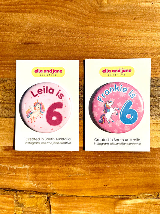Two unicorn birthday badges. The first unicorn party badge is light pink with darker pink hearts as the background. There is a flying unicorn with pastel rainbow tail and mane. The text is Leila is 6. The second badge is medium pink with lighter pink flowers as the background. There is a unicorn with darker rainbow tail and mane. The text is in blue and says Frankie is 6. The badges are on white product cards and laying on a piece of wood.