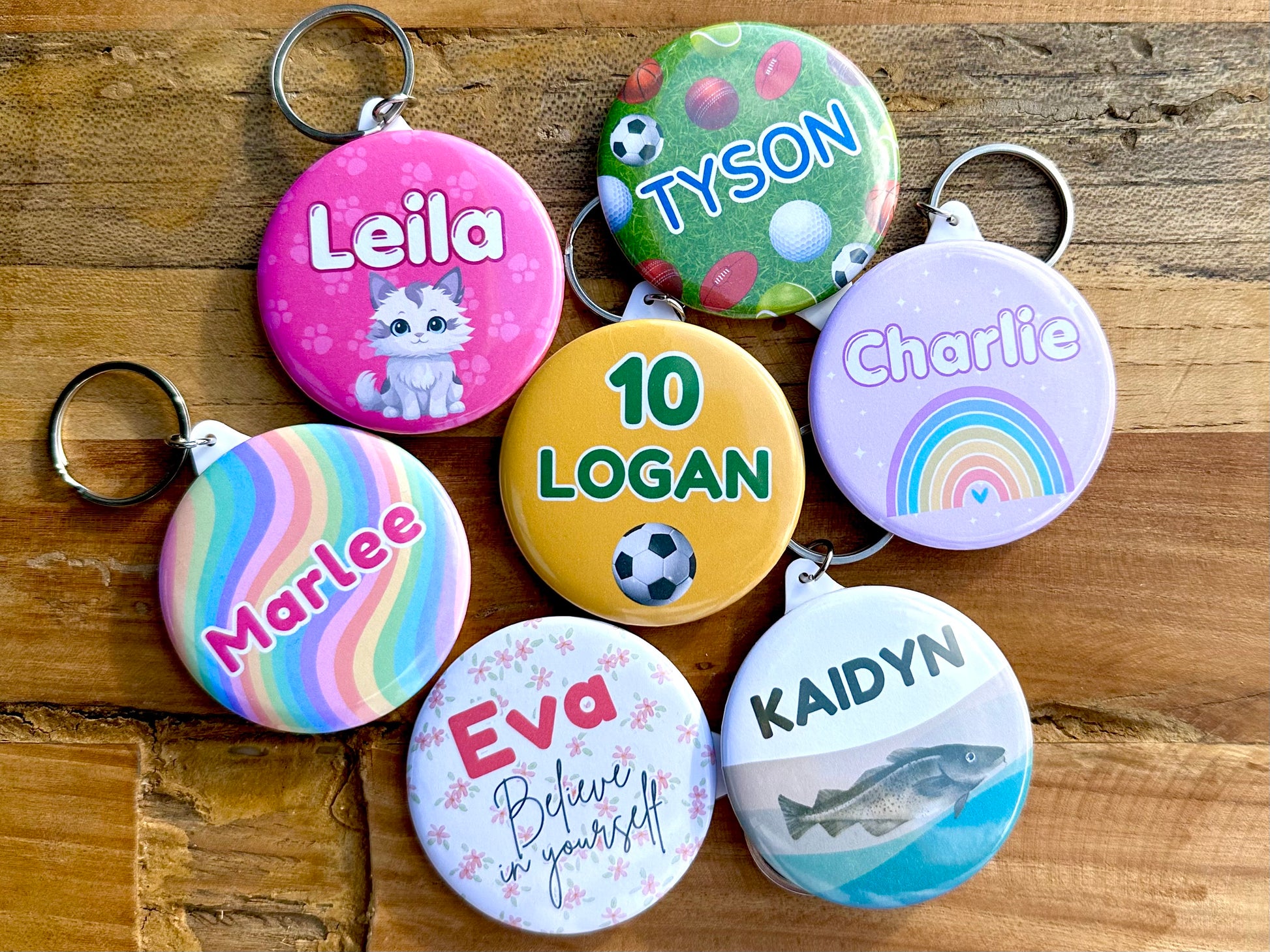 7 personalised keyrings with a name and image. One has a pink background with paw prints and a white and grey cat. One has green lawn and various sporting balls. One has a light purple background with a colourful rainbow. One has white grey and grey waves with a murray cod fish. One has a white background with soft pink flowers. One has a rainbow swirl background.