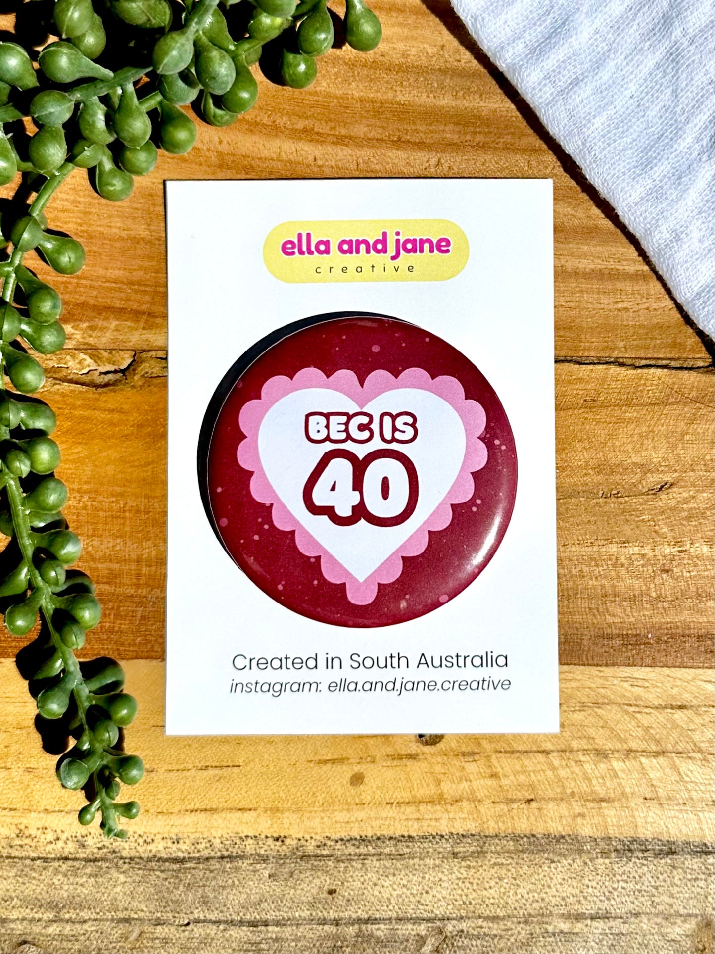 Pink and red hearts birthday badges. The party badge is a deep red maroon colour with light pink spots. There is a white heart in the centre with pink bubble ripples around the edge of the heart. There is text 'Bec is 40' in the centre of the white heart.  The birthday badge is sitting on white product cards and laying on a piece of wood with decorations.