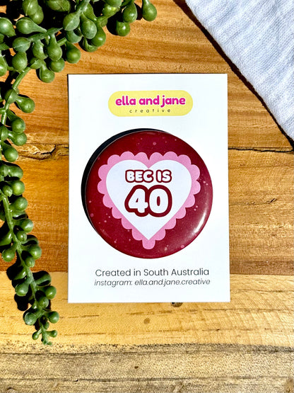 Pink and red hearts birthday badges. The party badge is a deep red maroon colour with light pink spots. There is a white heart in the centre with pink bubble ripples around the edge of the heart. There is text 'Bec is 40' in the centre of the white heart.  The birthday badge is sitting on white product cards and laying on a piece of wood with decorations.